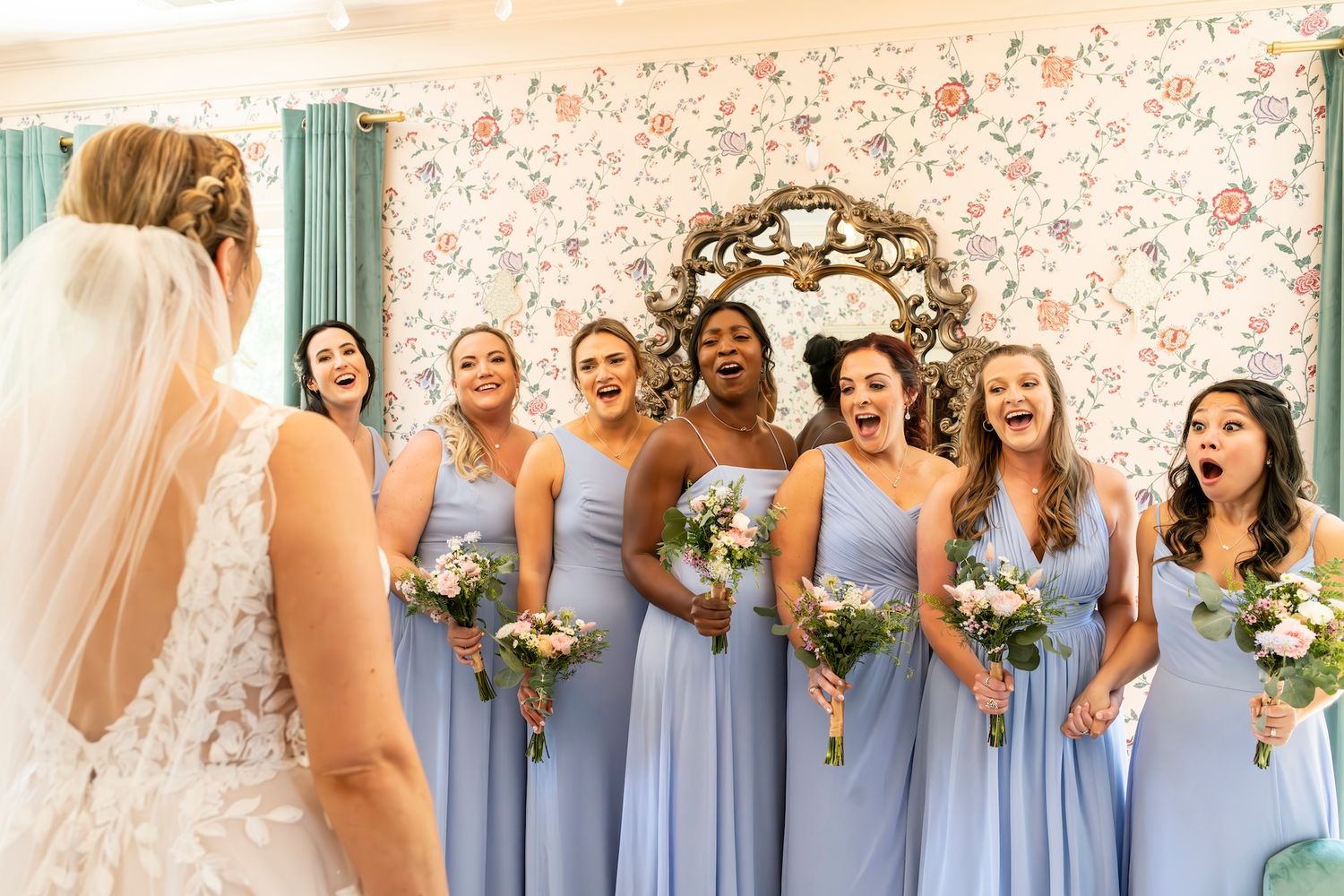 Bridesmaids in matching light blue dresses react joyfully to bride in white gown against floral wallpaper backdrop.