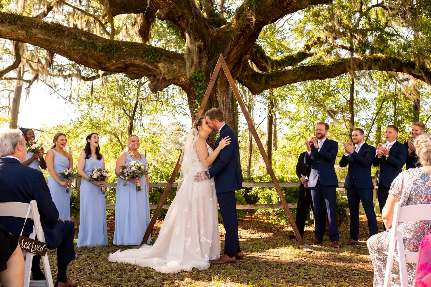 A couple shares a romantic kiss during their outdoor wedding ceremony under a majestic oak tree with Spanish moss.