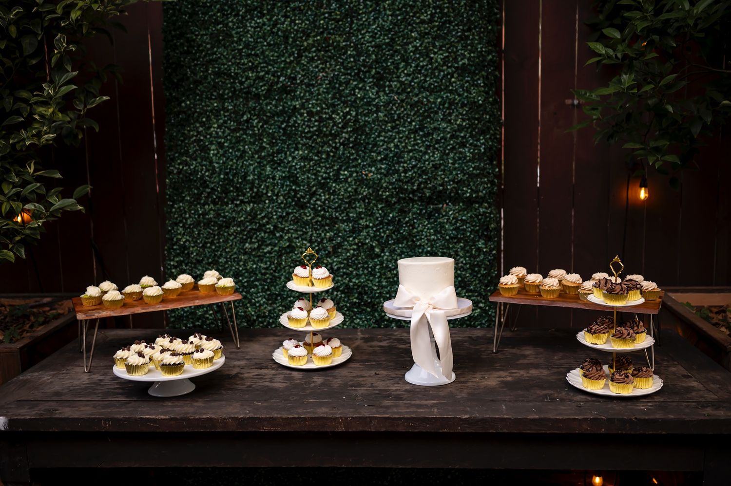 An elegant dessert table display with multiple tiers of cupcakes and a white cake stand against a dark green hedge backdrop.