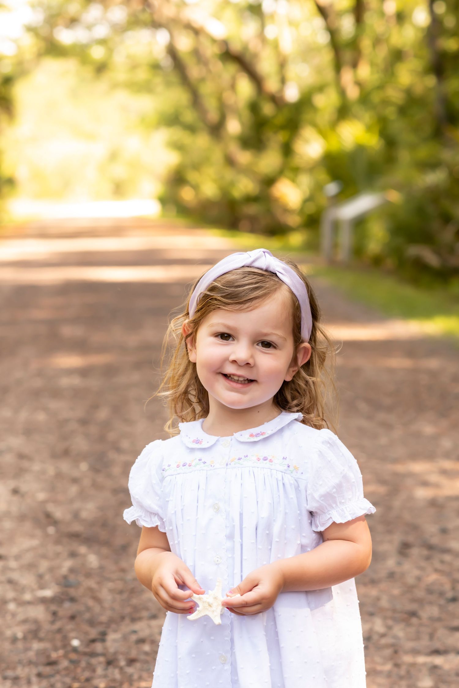 A small child wearing a white dress stands on a sunlit dirt path during a portrait session in nature.