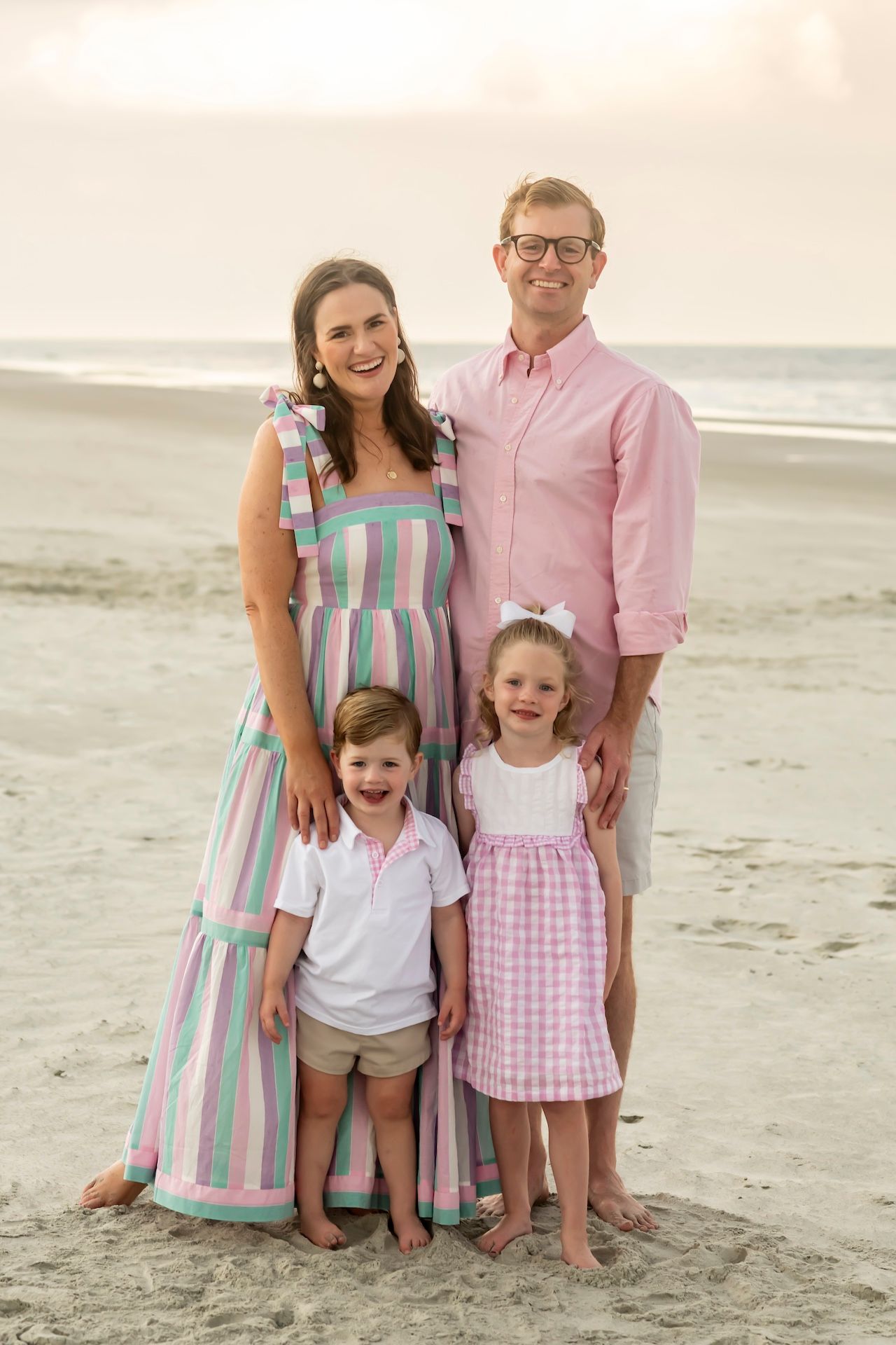 Family wearing coordinated pastel outfits pose together on sandy beach during sunset.