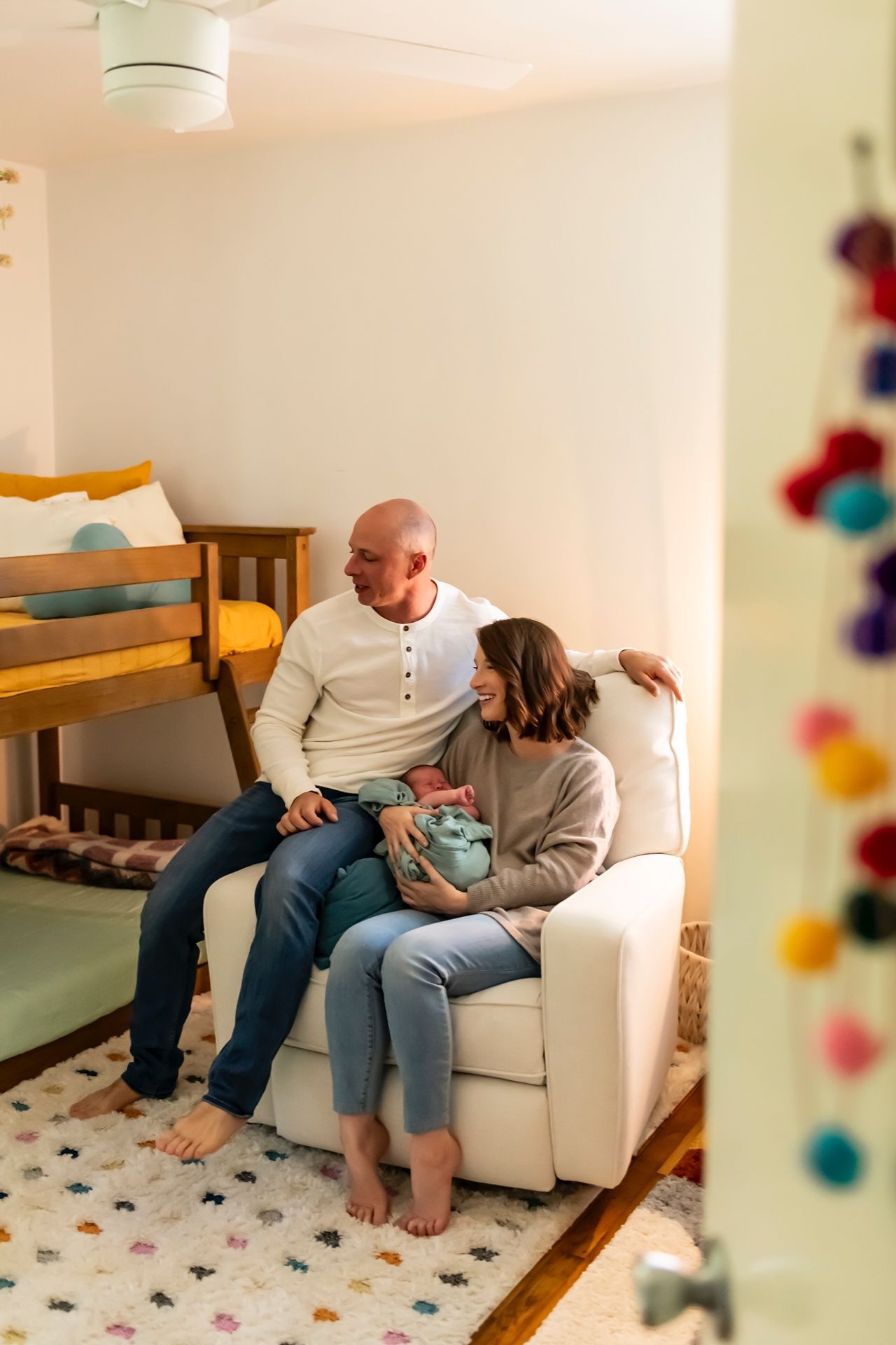 Two people sit together on a white armchair in a cozy nursery room with a colorful pom pom garland and bunk bed.