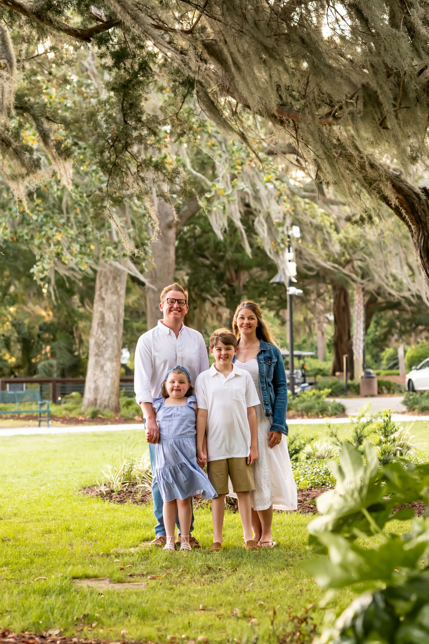 A family stands together on a lush lawn under Spanish moss-draped oak trees in a serene park setting.