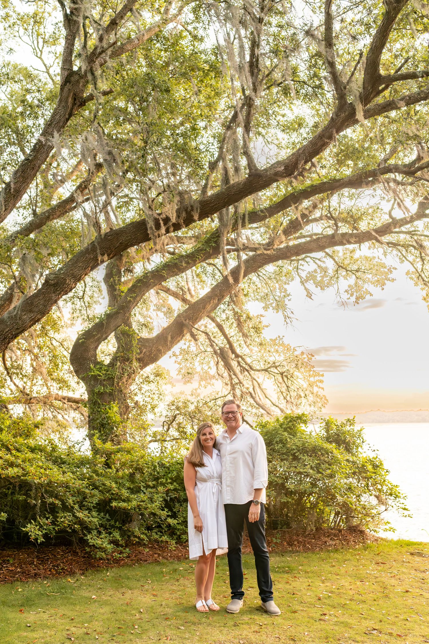 Two people stand together on a grassy lawn beneath a sprawling oak tree with Spanish moss at sunset by the water.