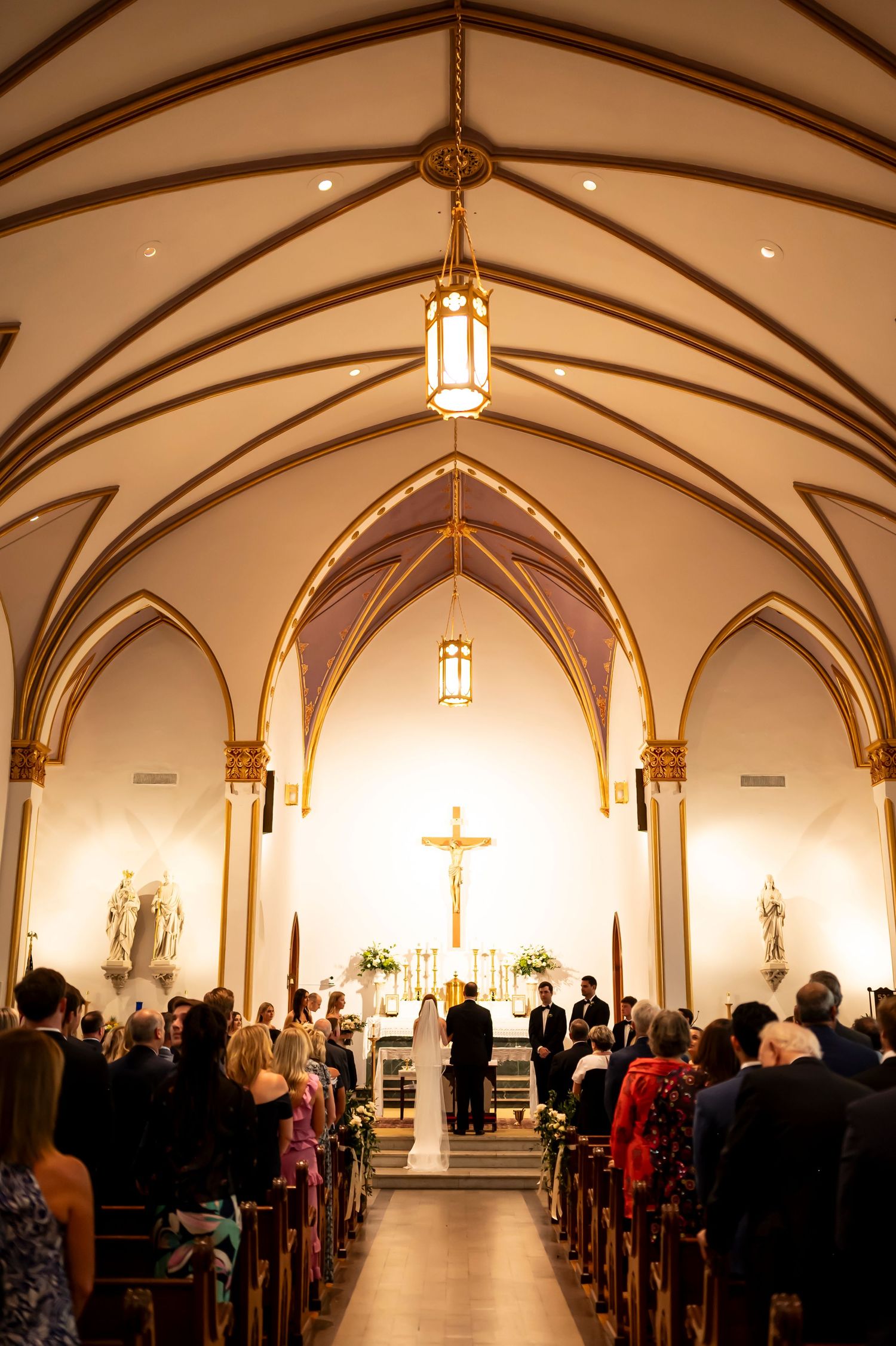 A Catholic church wedding ceremony in a Gothic-style chapel with vaulted ceilings and hanging lanterns illuminating the aisle.