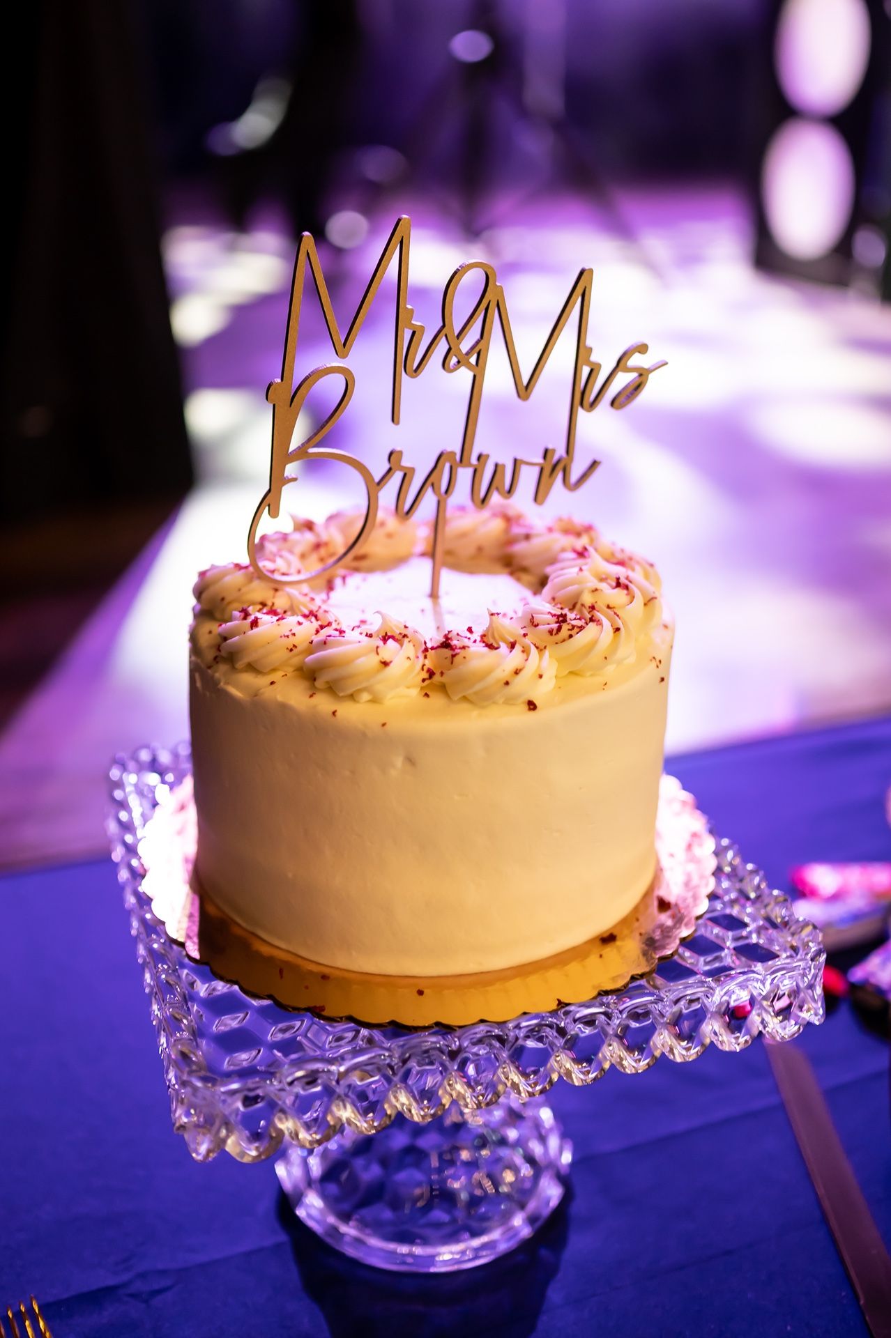 Elegant wedding cake with gold 'Mr & Mrs Brown' topper displayed on crystal stand with purple lighting in background.