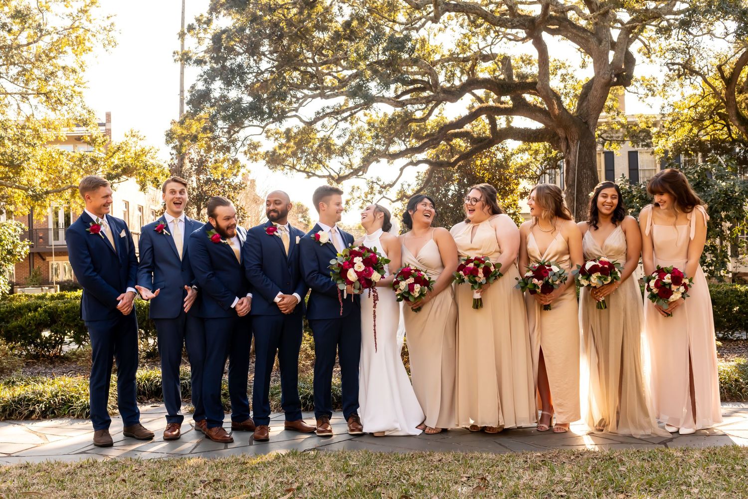 Wedding party in navy suits and blush bridesmaid dresses pose together outdoors under a large oak tree at sunset.