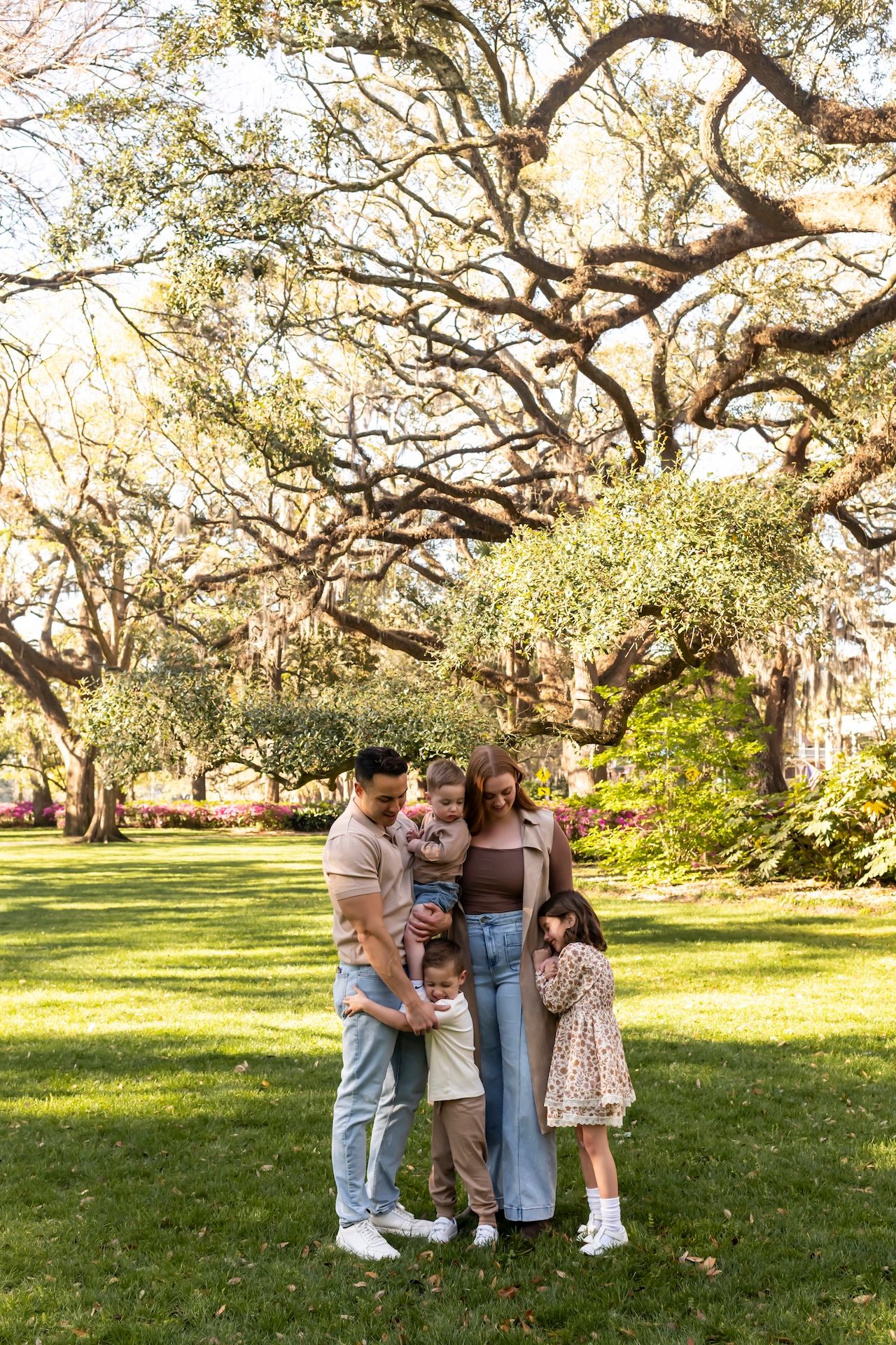 A family stands together under sprawling oak trees with Spanish moss on a sunny day in a lush park setting.