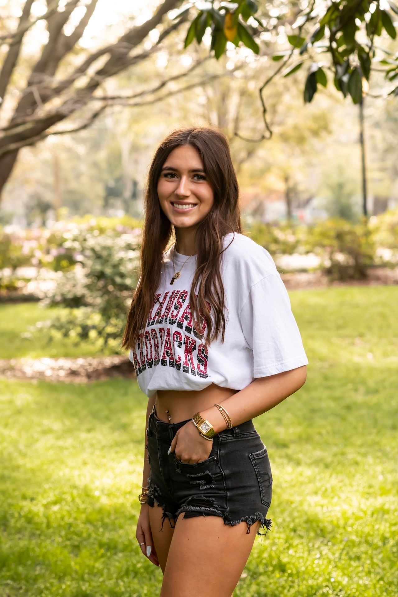Person in a white Arkansas t-shirt and denim shorts poses outdoors in a sunny park setting.