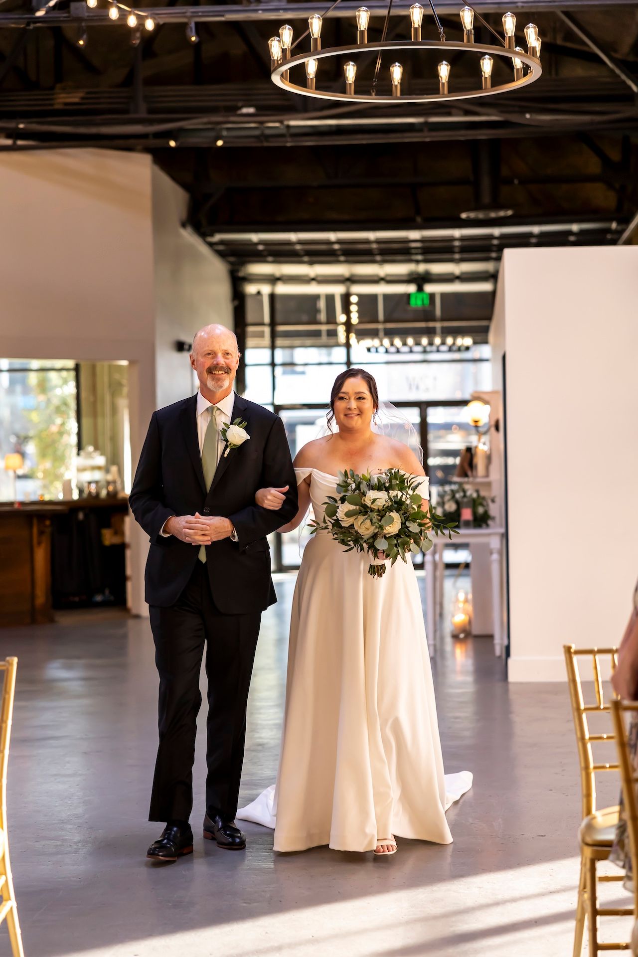 A formal wedding processional in an industrial-modern venue with circular chandelier and natural lighting.