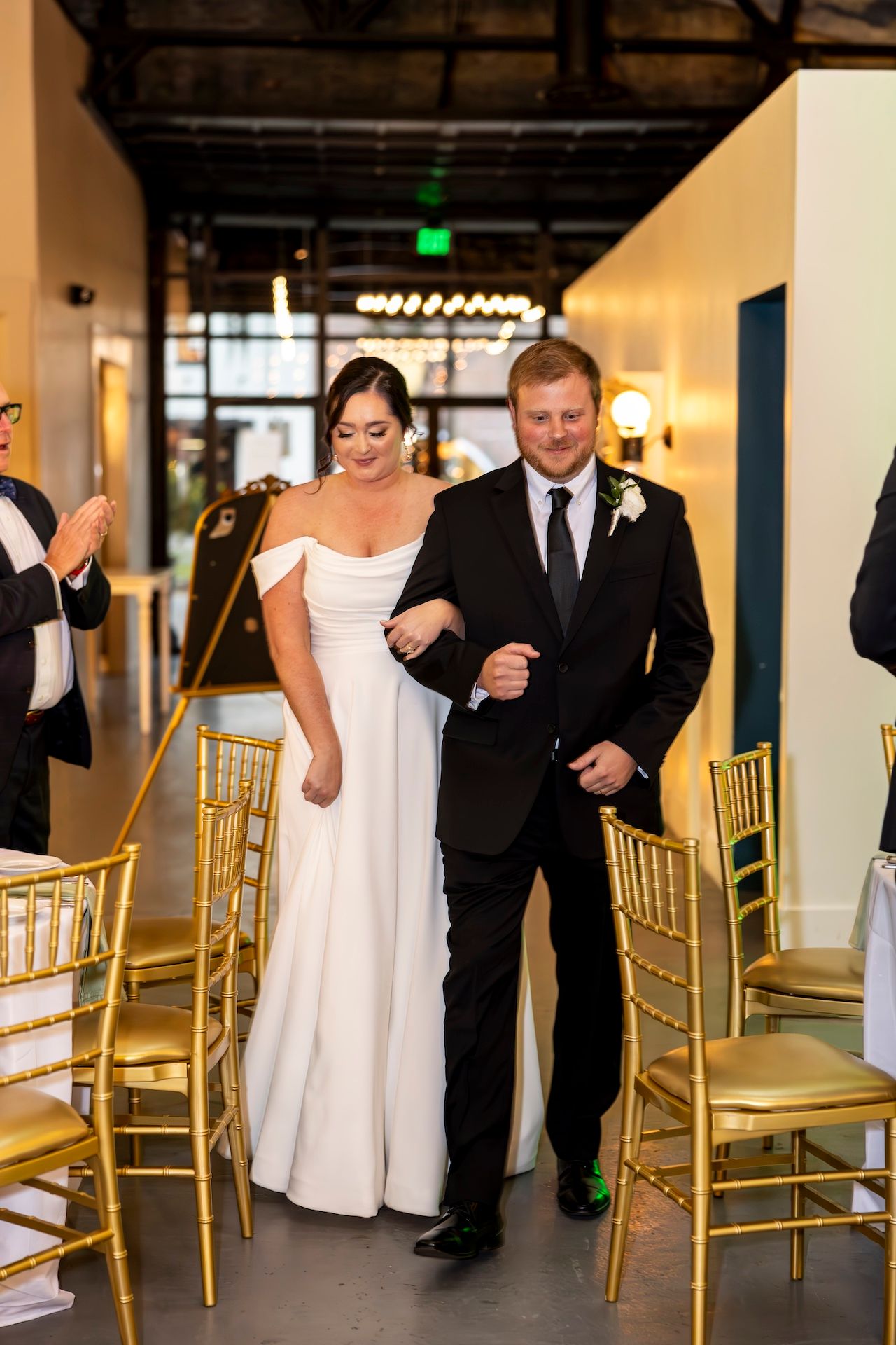 A person in an elegant white off-shoulder wedding gown walks down an aisle with an escort past gold chiavari chairs.
