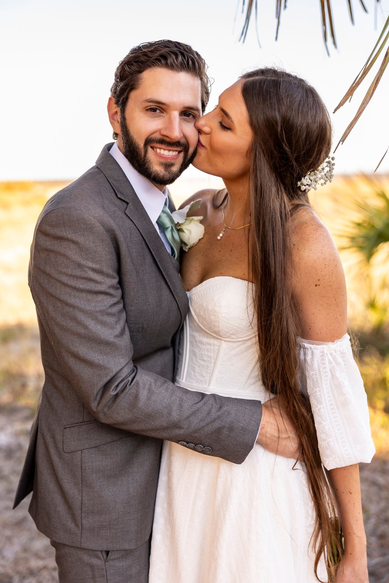 A newlywed couple share an intimate moment outdoors, with the bride wearing white and groom in grey suit.