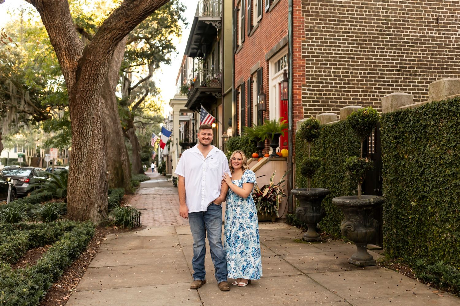 A couple walks together on a tree-lined sidewalk in front of historic brick townhouses on a sunny afternoon.