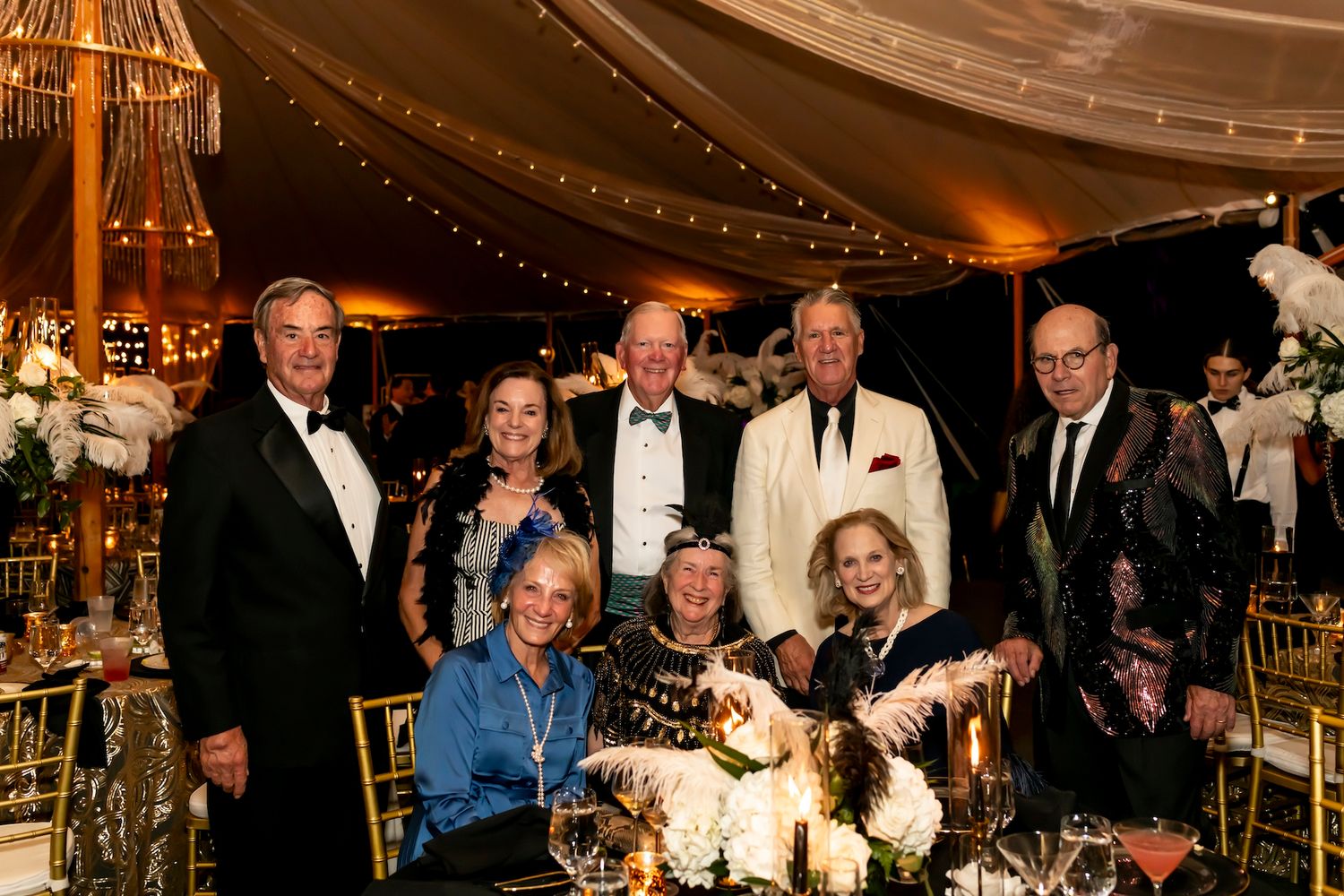 A formal evening event with guests gathered around an elegantly decorated table with floral centerpieces and ambient lighting.