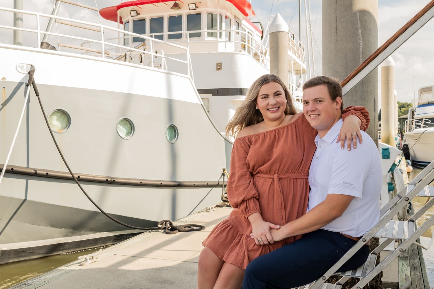 A couple sits together and smiles while posing by a docked boat in a marina on a sunny day.