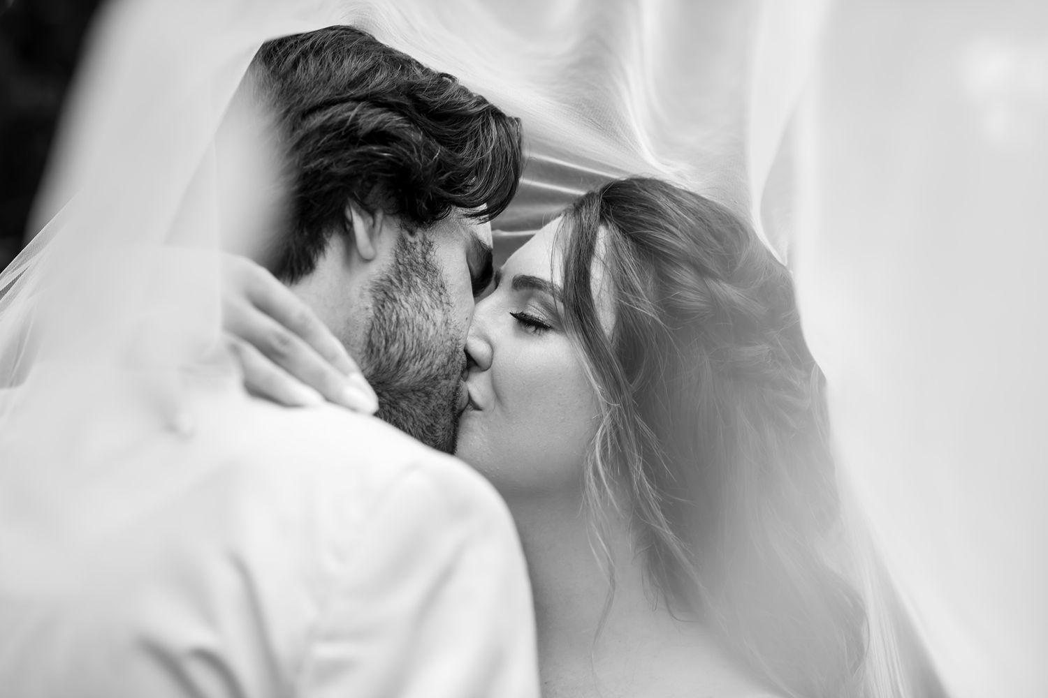 Black and white photograph of romantic wedding kiss with bride wearing traditional white veil and gown.