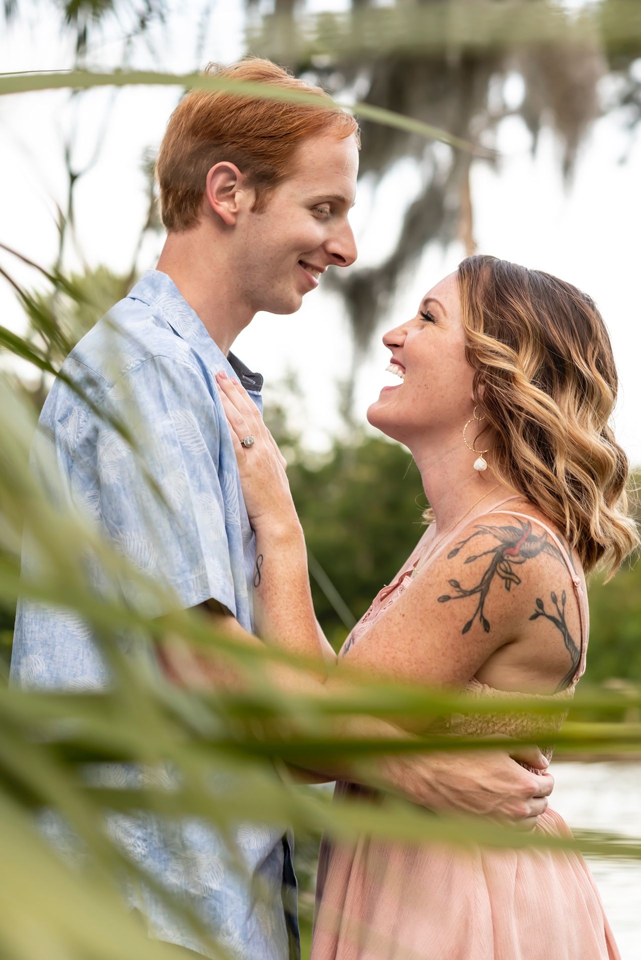 A romantic couple embraces and smiles at each other outdoors, with palm trees visible in the background.