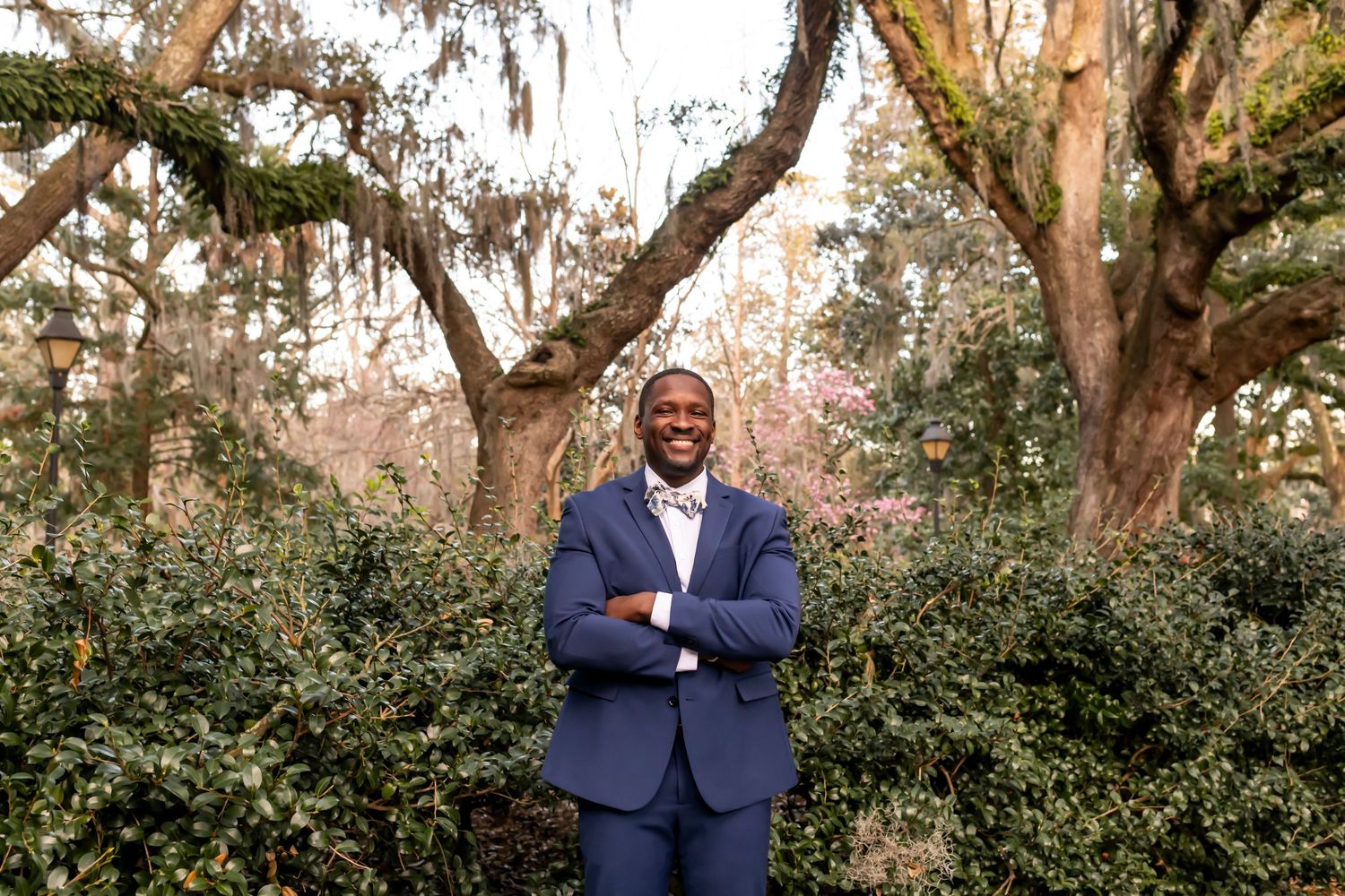 A confident person in a navy business suit stands with crossed arms in a lush garden with Spanish moss draped trees.