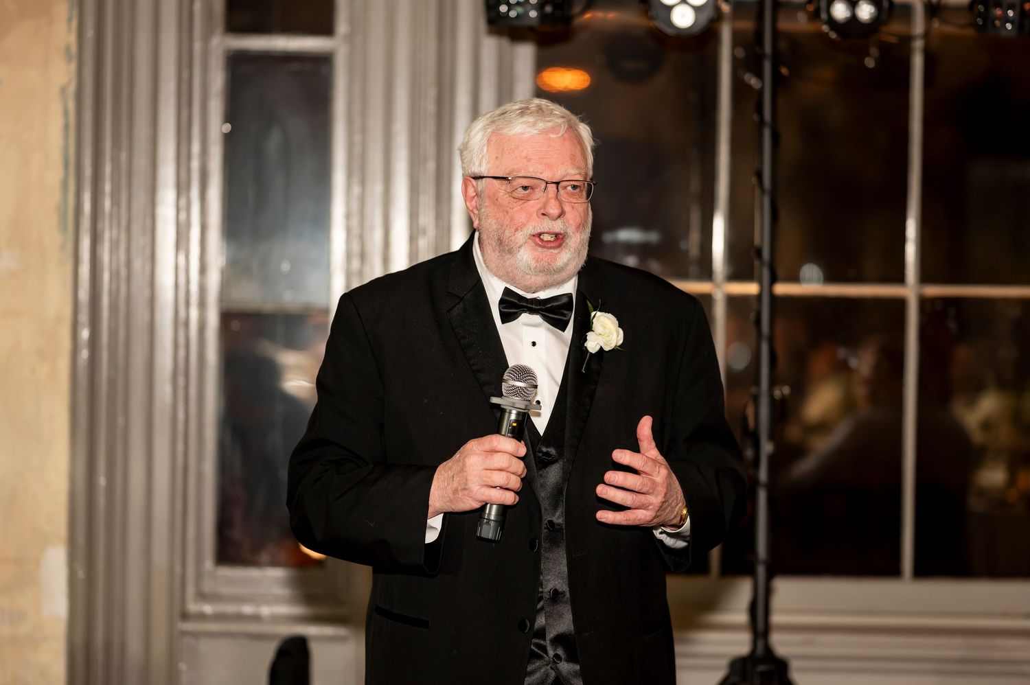 A person in a black tuxedo gives a speech while holding a glass at an elegant indoor event.
