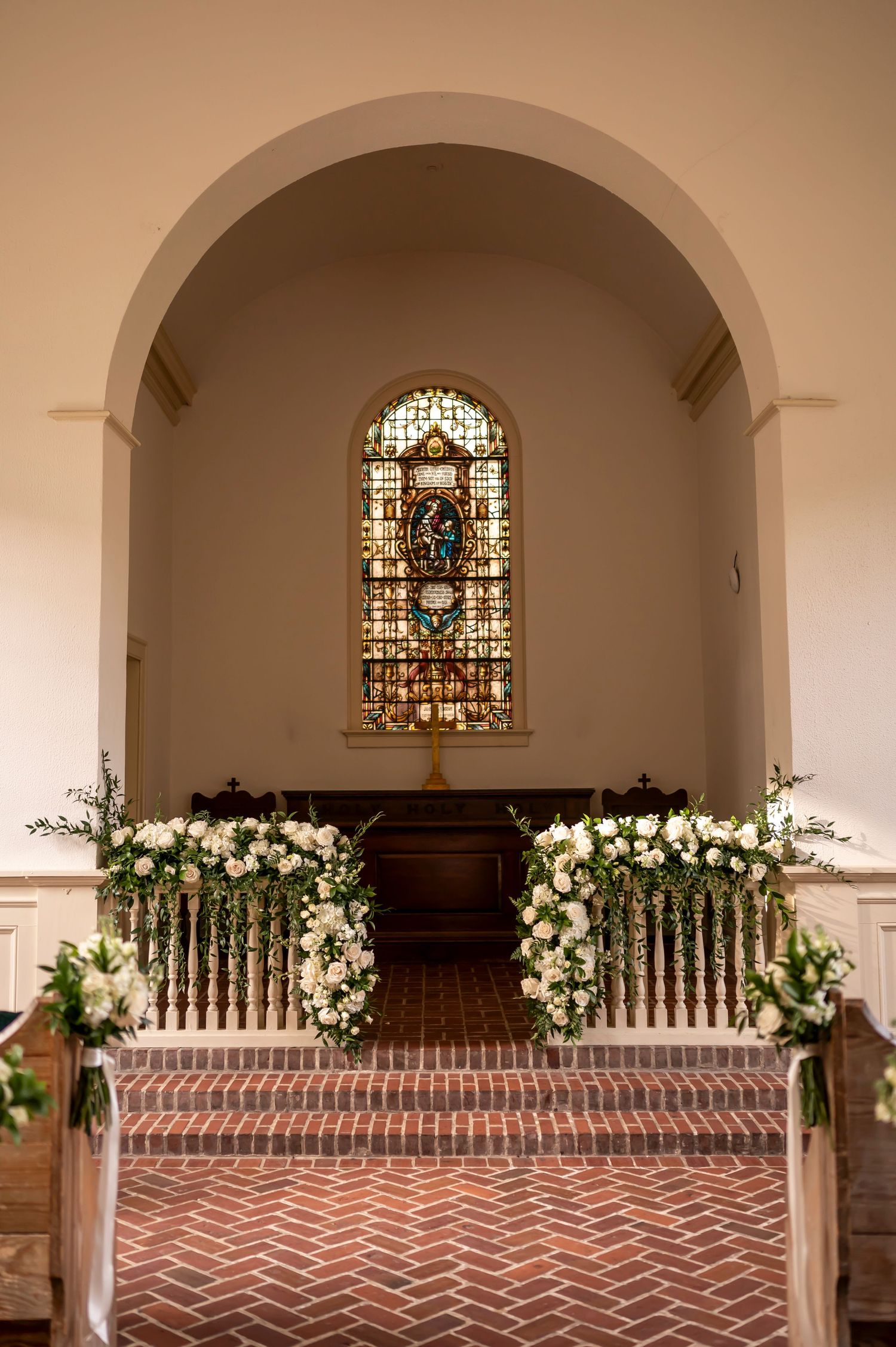 Elegant church altar with stained glass window and symmetrical white floral arrangements on brick steps beneath an arched entryway.