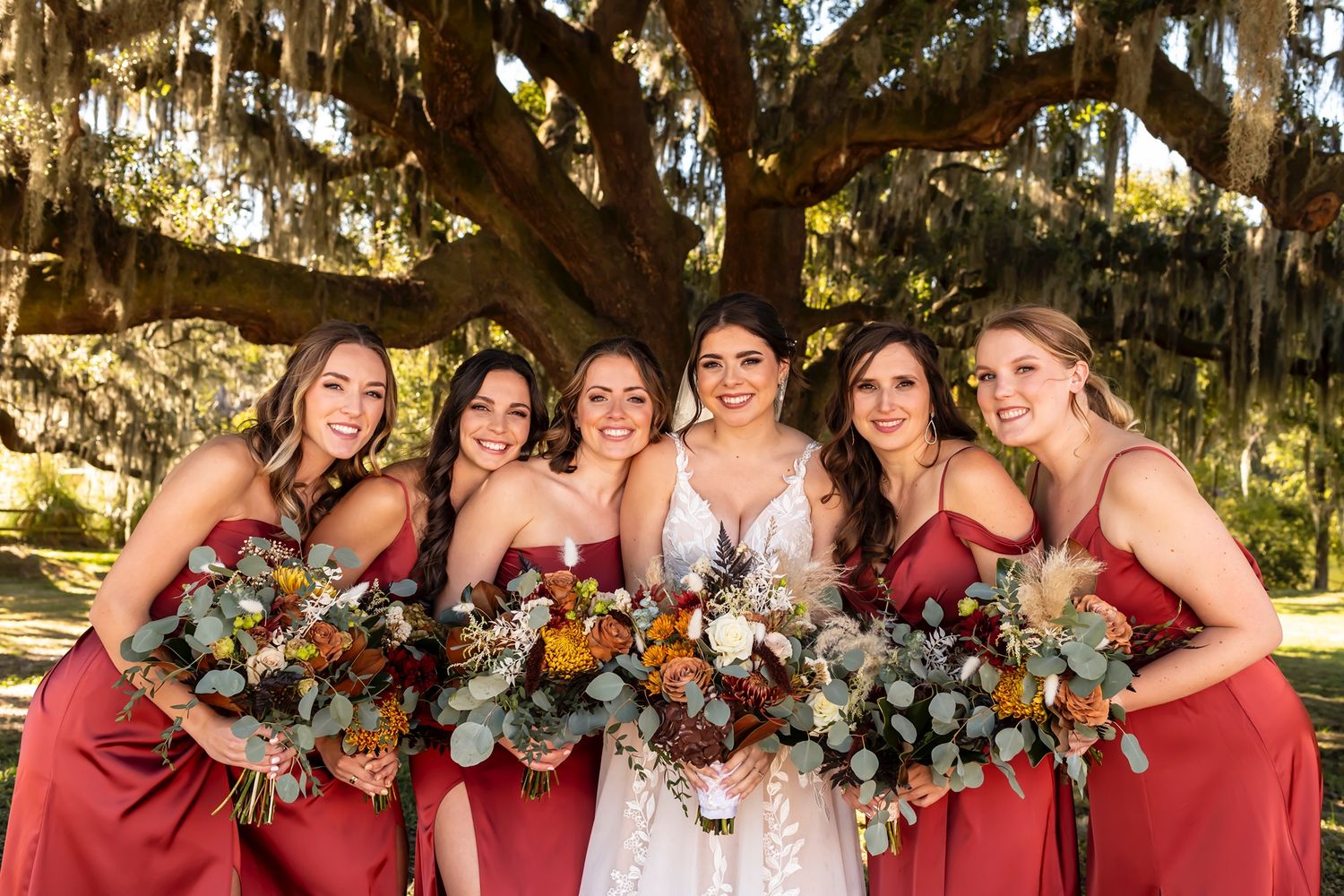 Wedding party in burgundy dresses with rustic fall bouquets pose under Spanish moss draped oak tree.