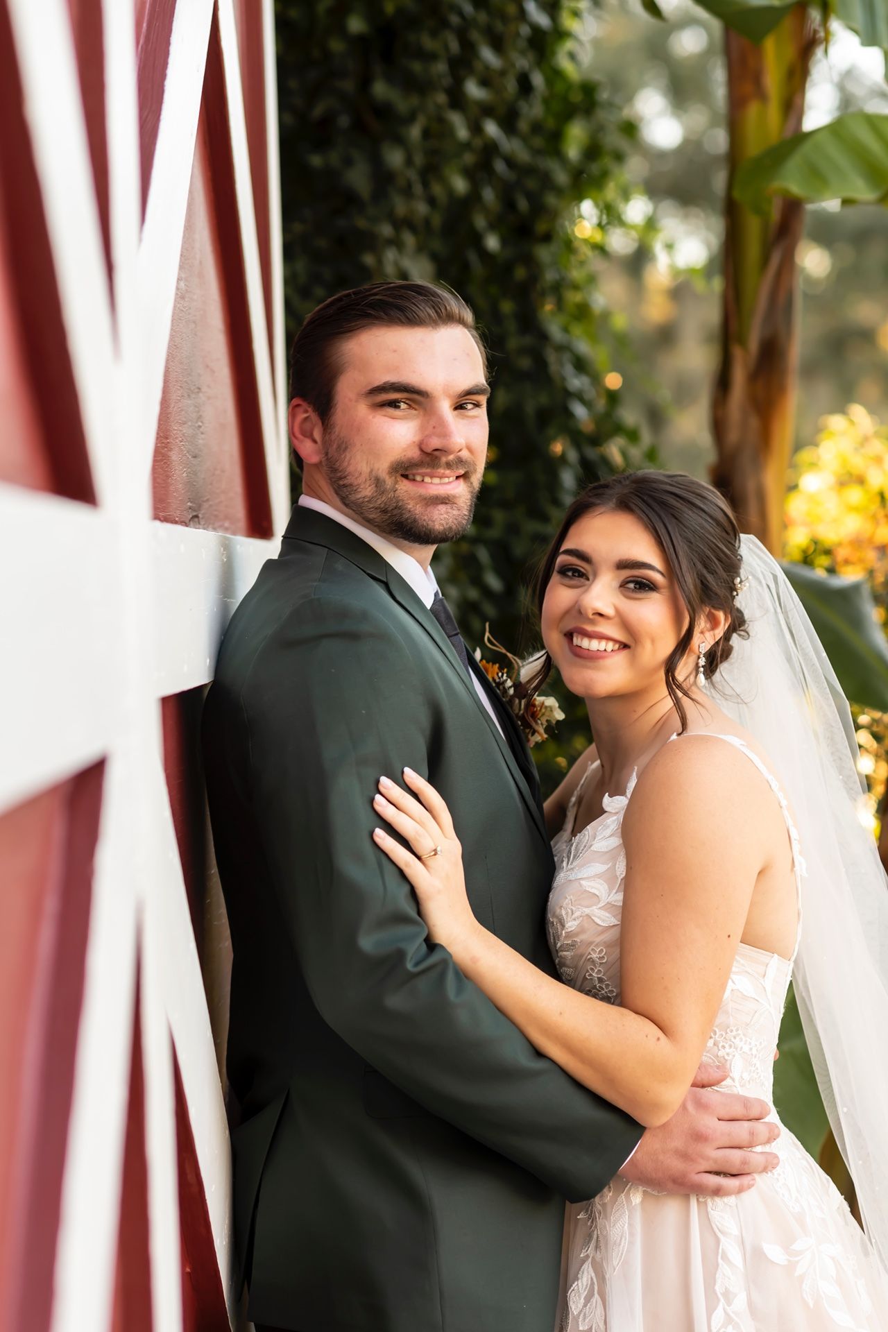 A newlywed couple pose together outdoors against a red barn wall with greenery and natural lighting.