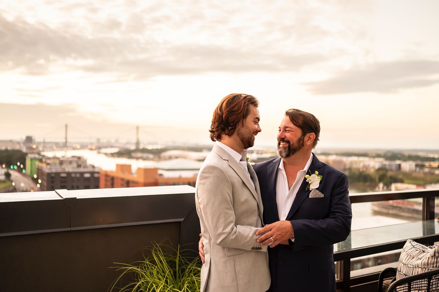Two people in formal attire share a romantic moment on a rooftop terrace overlooking a cityscape at sunset.