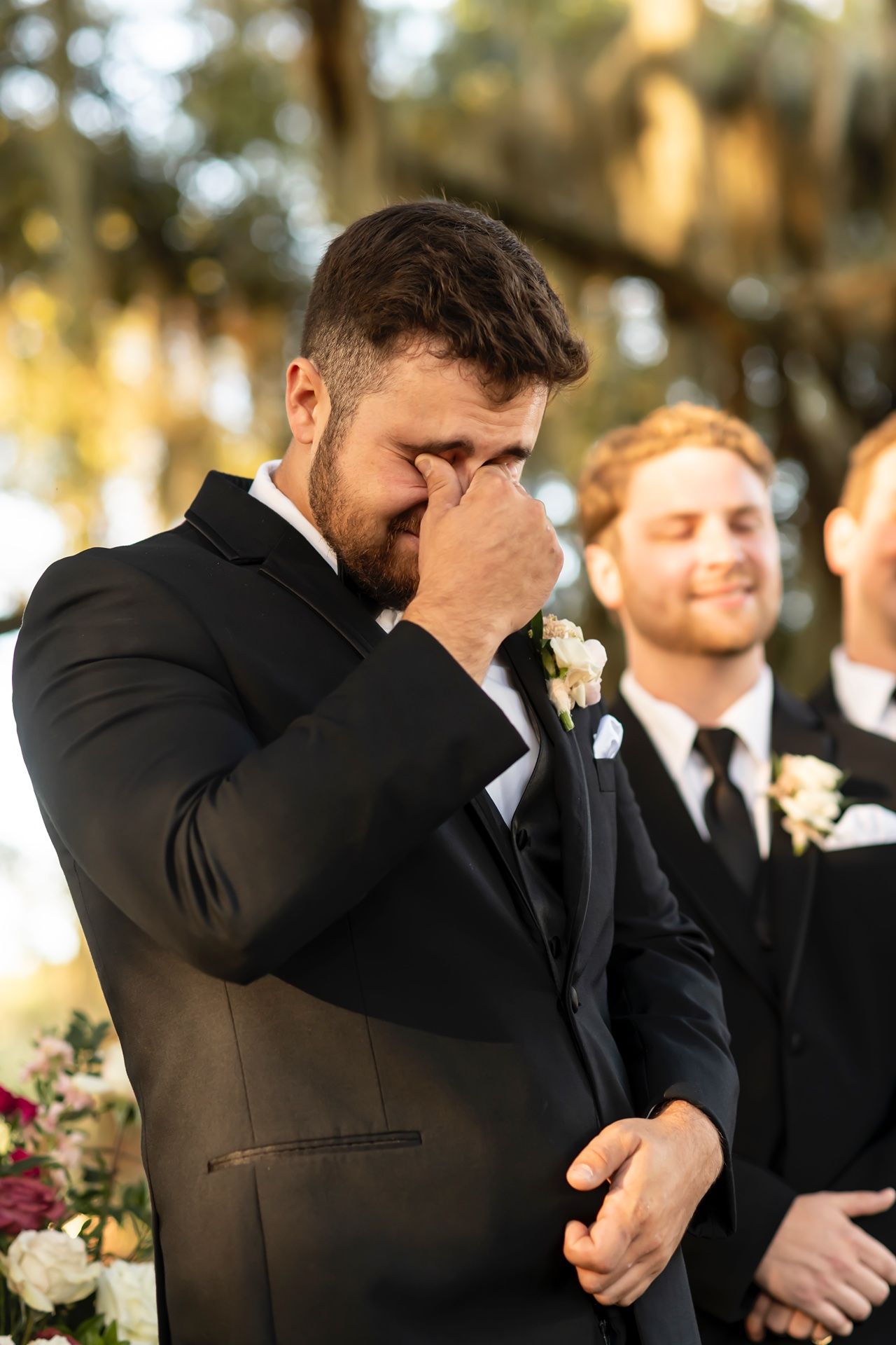 Emotional groom wipes away tears during outdoor wedding ceremony while wearing a black tuxedo.