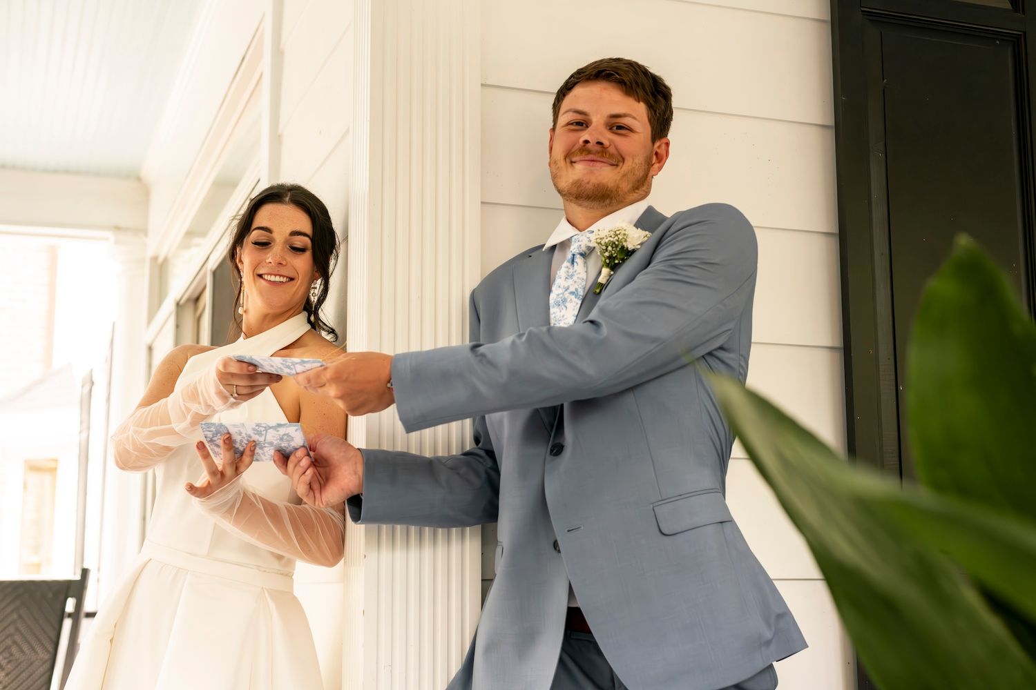 Newlyweds stand on a white porch as they hold up their arms showing off rings during outdoor wedding photography.