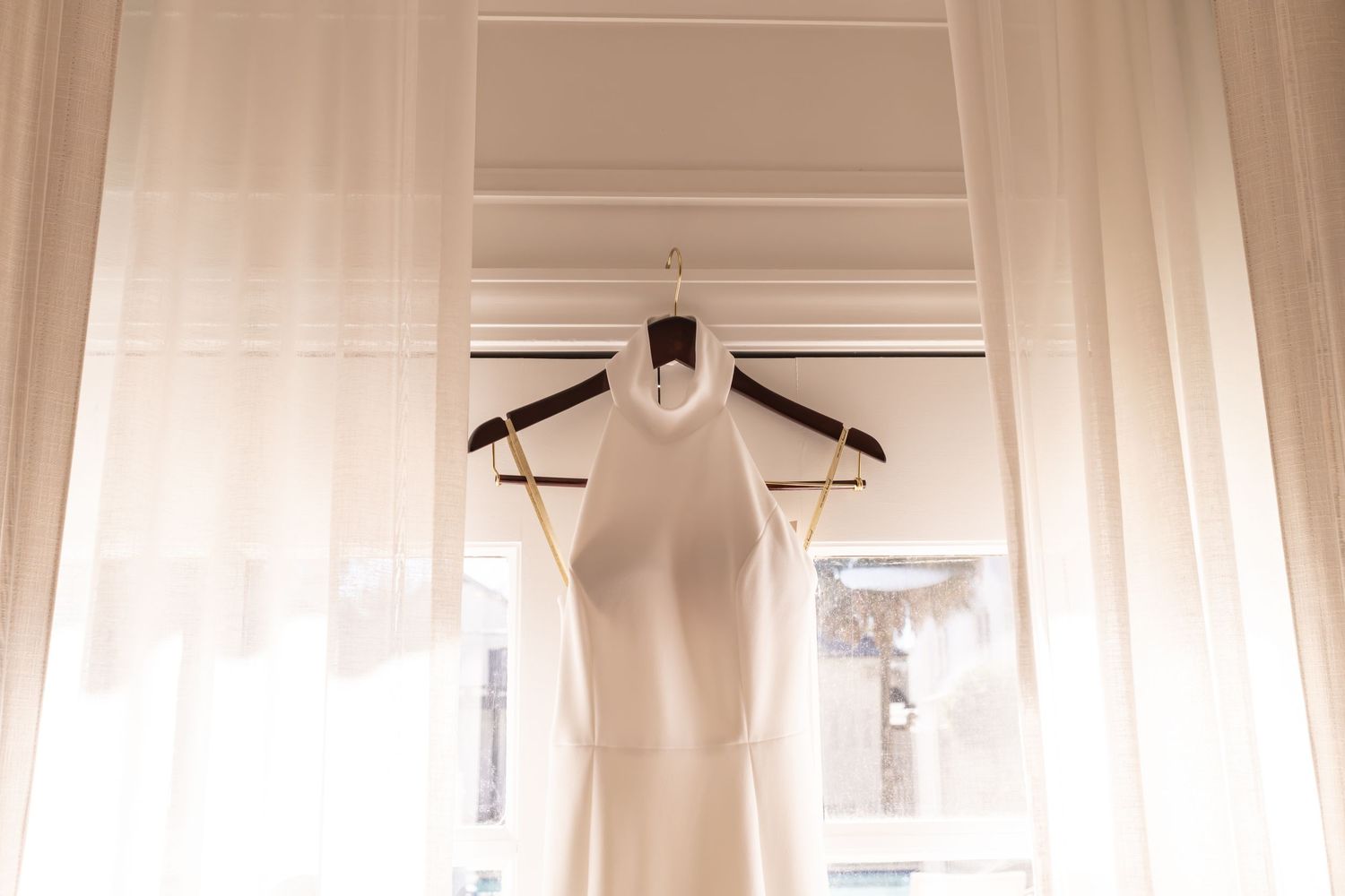 A white wedding dress hangs elegantly on a wooden hanger near a window with sheer curtains creating a soft ethereal glow.