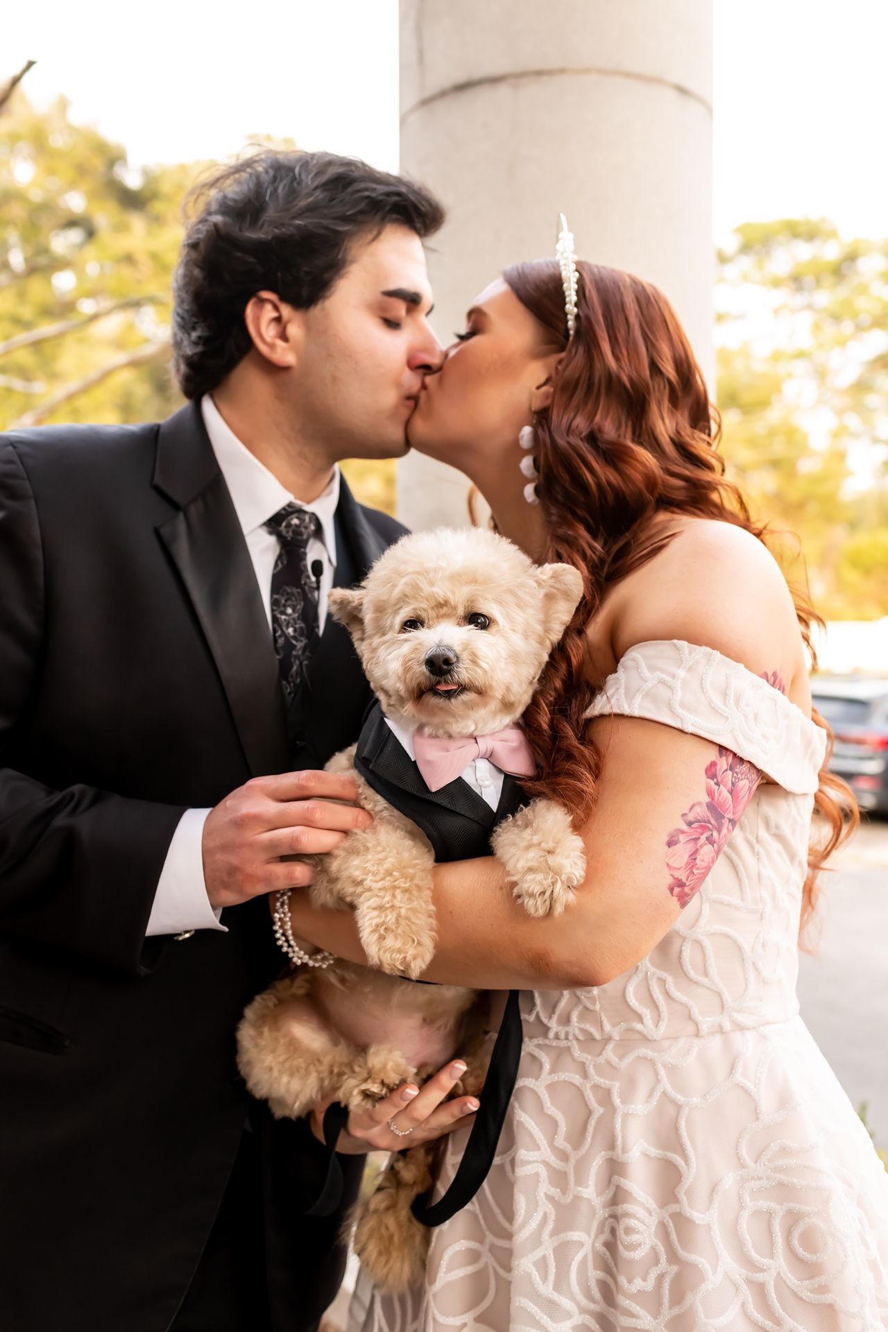 A couple shares a wedding kiss while holding their small fluffy dog dressed in formal attire.