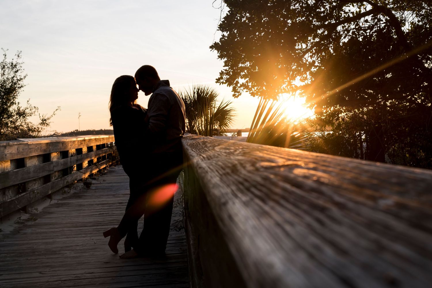 Silhouetted couple embracing on wooden boardwalk at sunset with sun flare and coastal scenery in background.