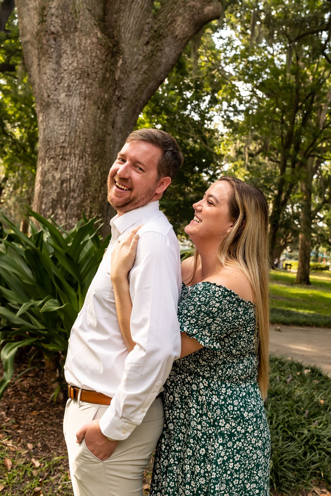 A couple shares a playful candid moment outdoors in a park, with the woman in a floral dress laughing behind her partner.