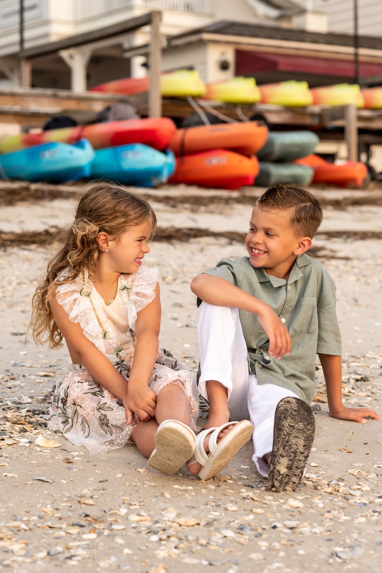 Two young children sitting together on a beach shore with colorful kayaks in the background on a sunny day.