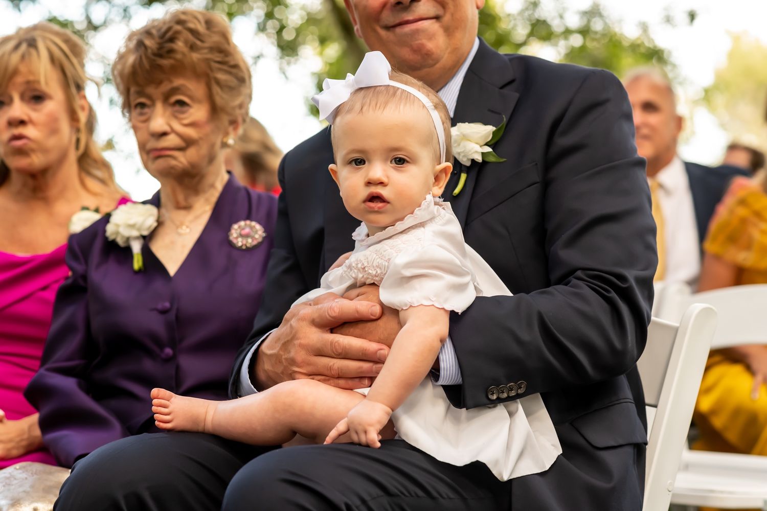 A small baby in white clothing sits with guests at an outdoor wedding ceremony.
