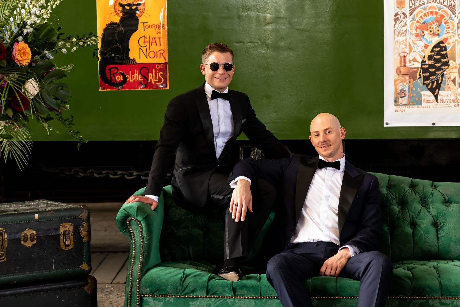 Two people in tuxedos sit on an emerald green velvet couch with vintage posters on the green wall behind them.