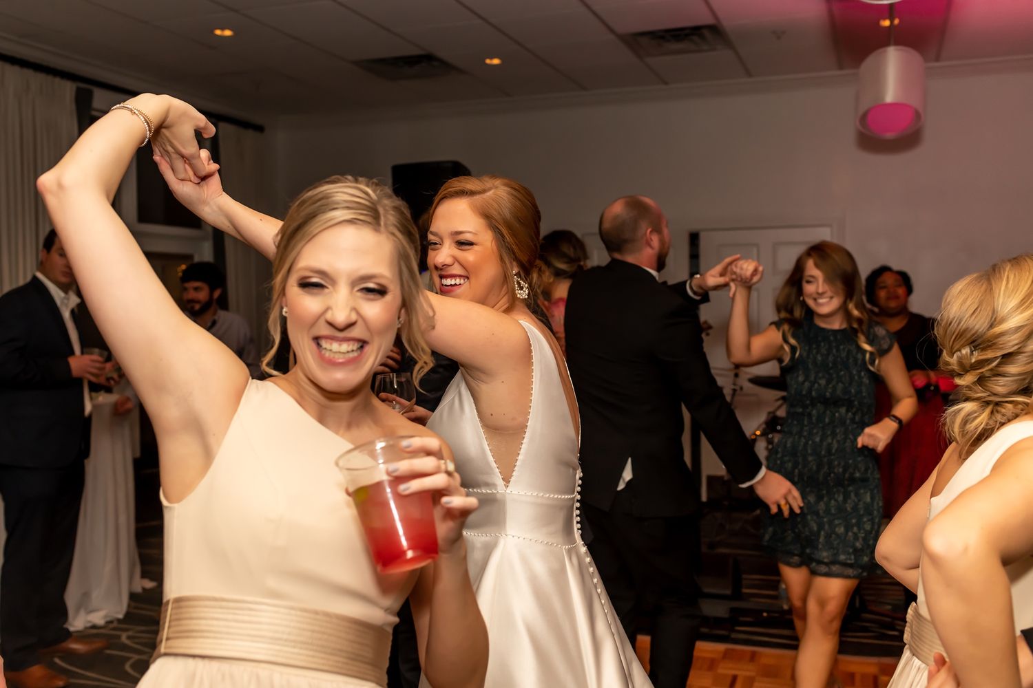 Wedding guests dancing and celebrating on the dance floor at a reception with pink lighting in the background.