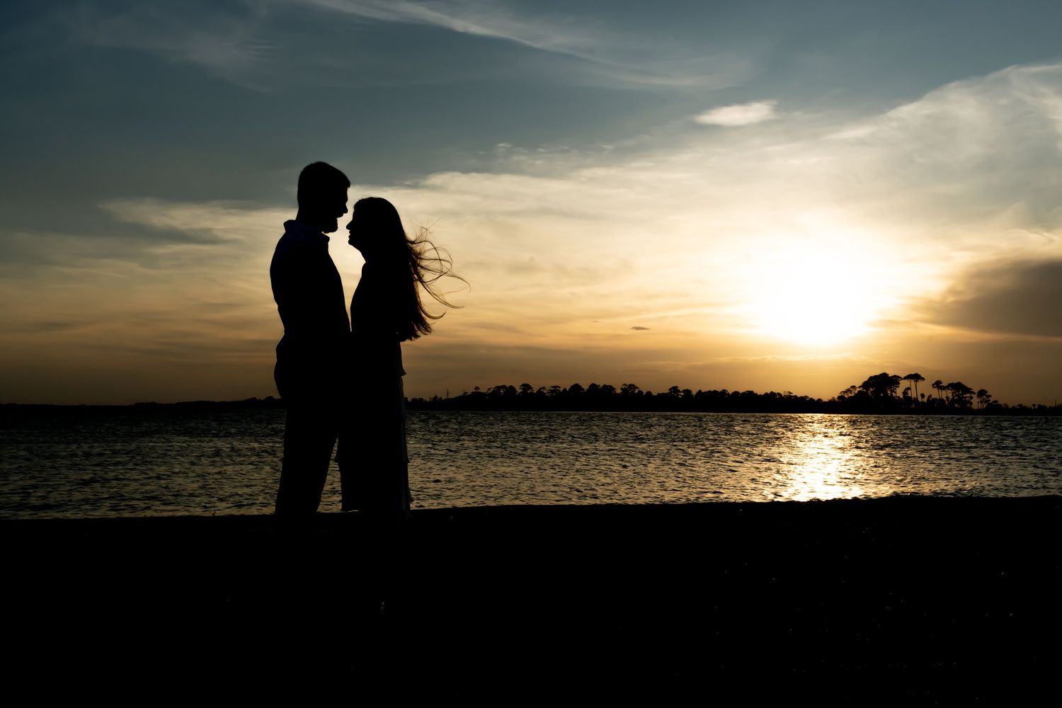 Silhouetted couple standing together against dramatic sunset over water with glowing orange sky.
