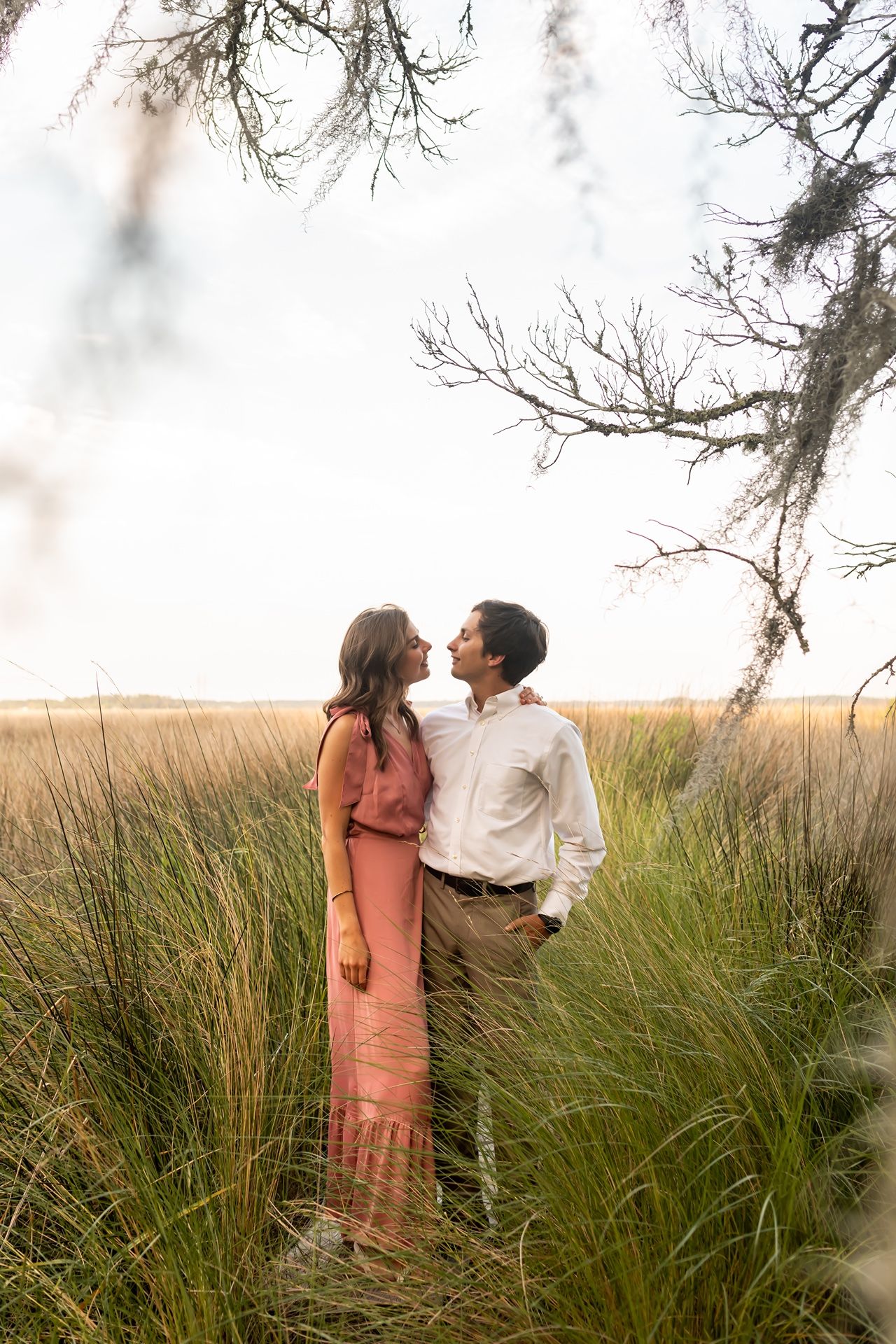 A romantic couple stands together in tall grass with bare tree branches overhead during golden hour.