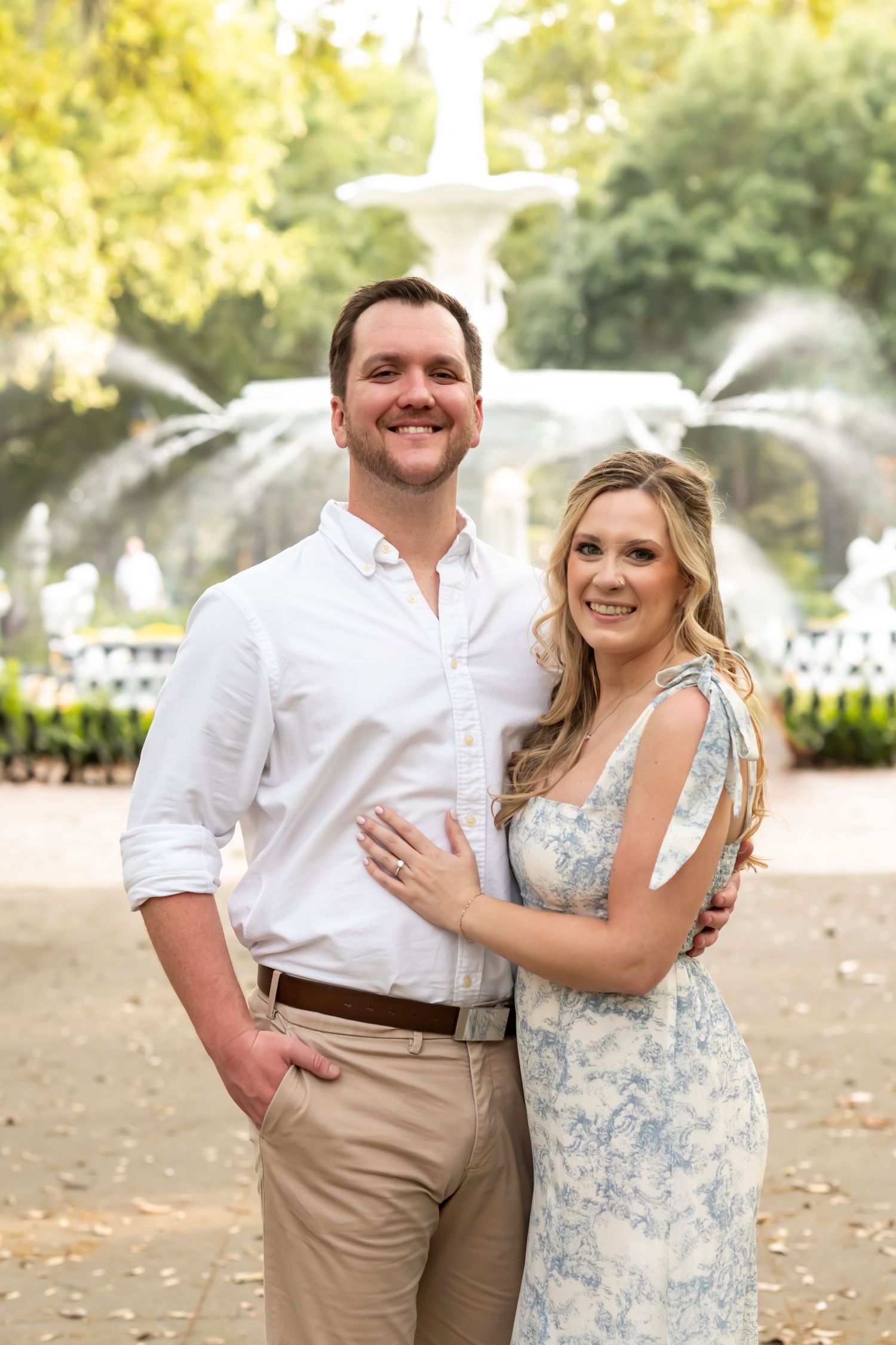 A couple poses together outdoors in front of a white fountain wearing coordinated light-colored outfits.