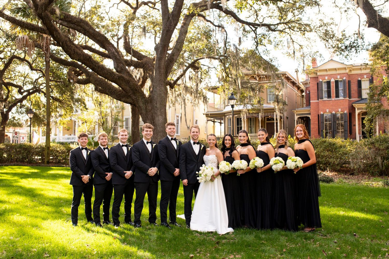 Wedding party in formal attire poses on a lawn with Spanish moss draped trees and historic buildings in background at Forsyth Park.