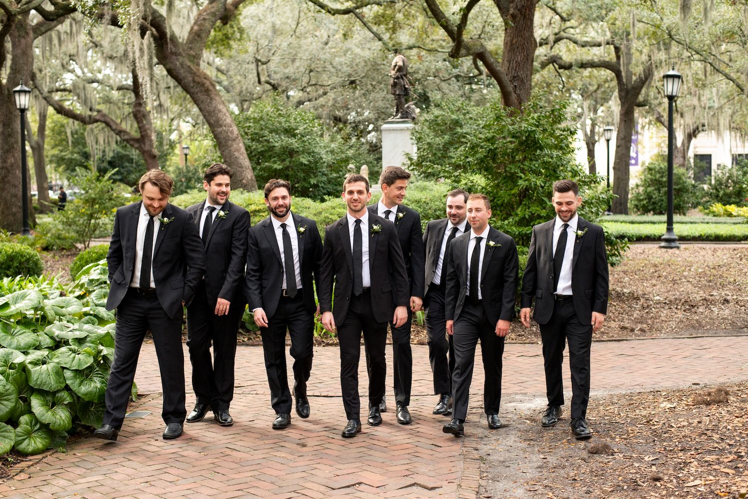 Groomsmen in black suits and ties walking together through a scenic outdoor garden path during a wedding photoshoot.