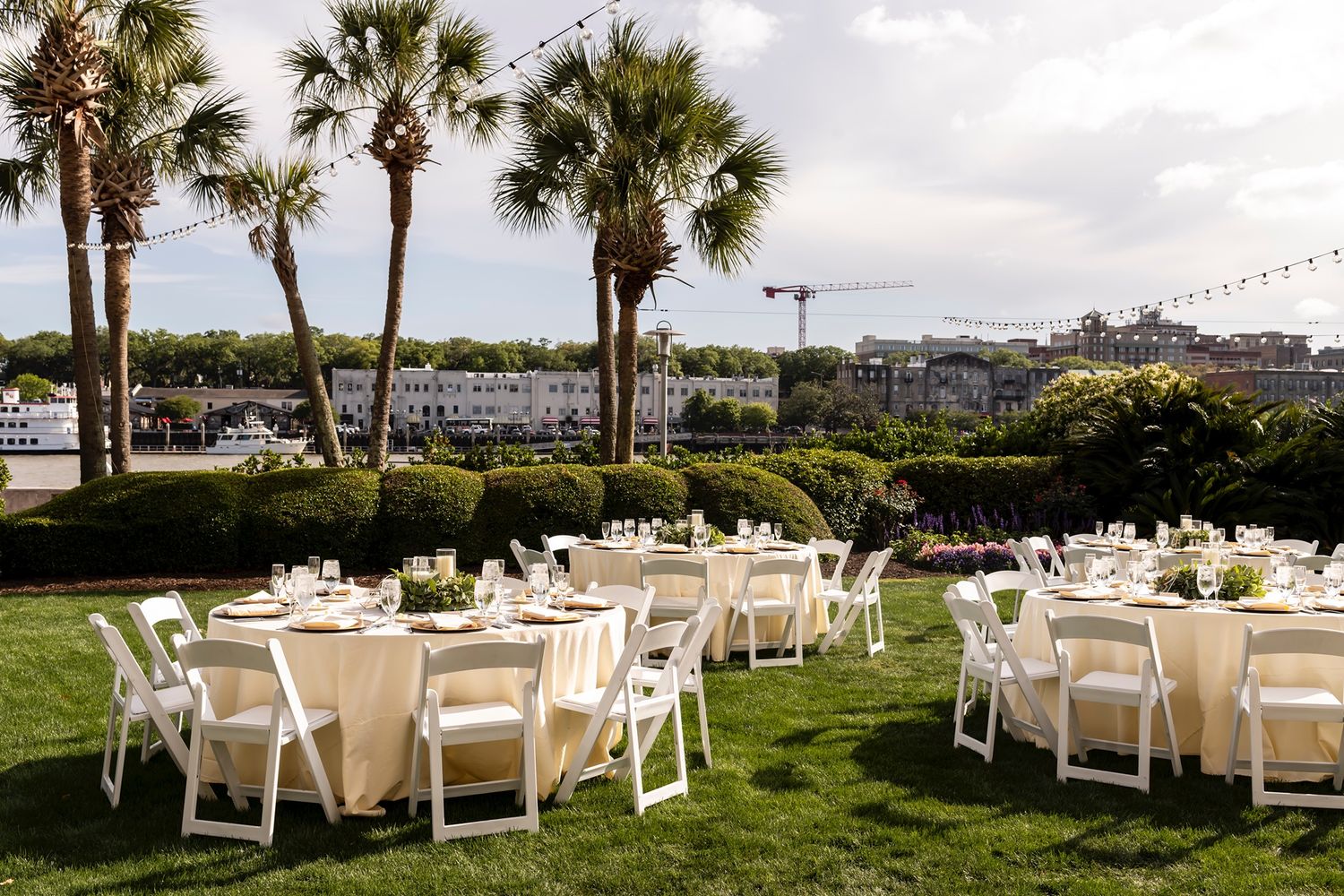 Outdoor wedding reception setup with round tables and white chairs on a lawn overlooking the marina with palm trees.