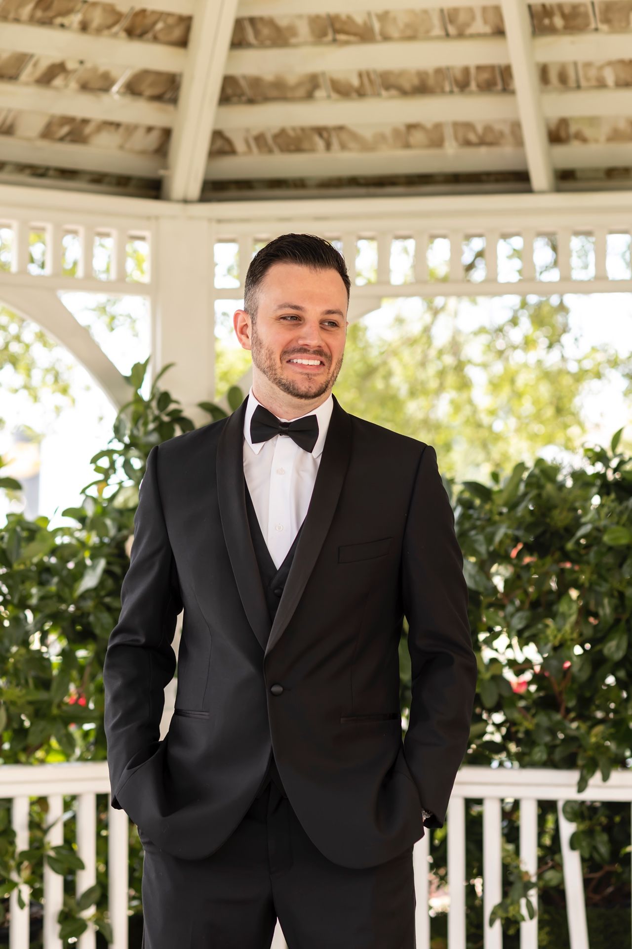 Well-dressed groom in black tuxedo and bow tie smiling while standing in white garden gazebo on sunny day.