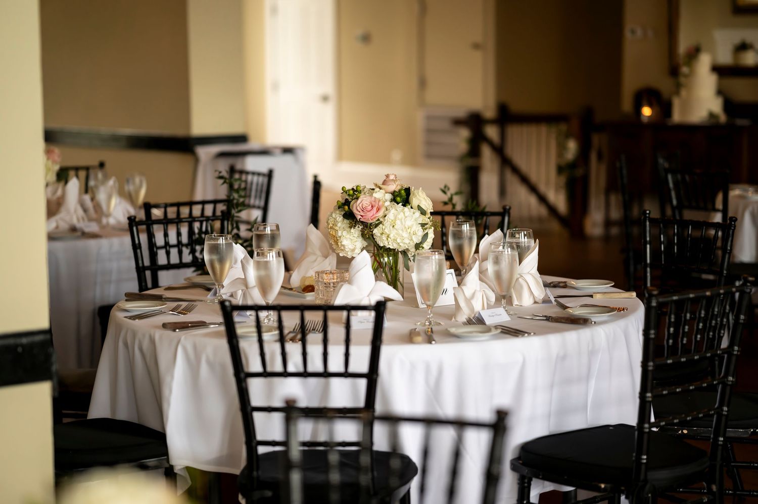 Elegant wedding reception venue with white linen covered round tables, black chiavari chairs, and floral centerpieces.