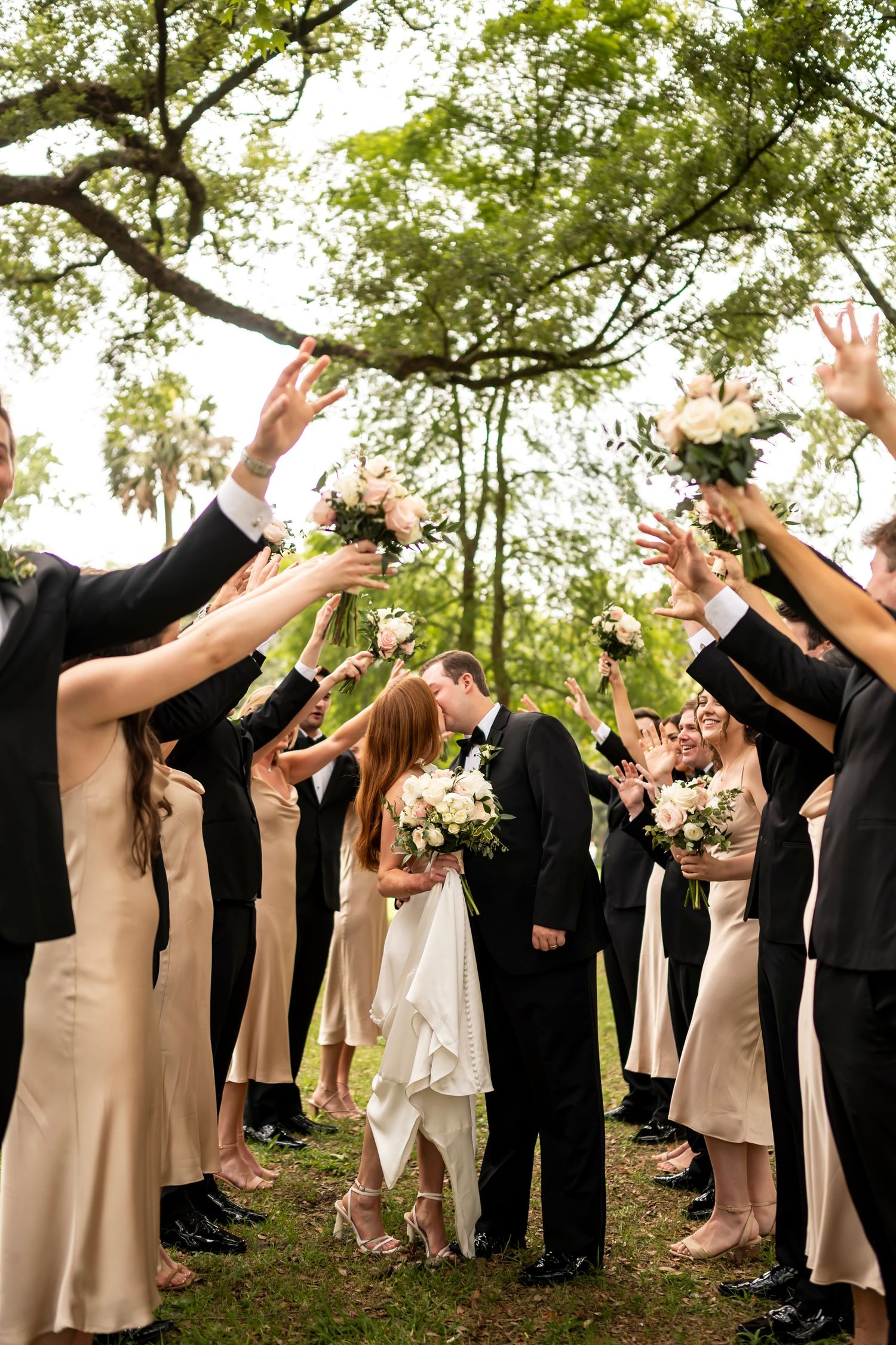 Wedding party in black suits and beige dresses raises arms joyfully under trees at outdoor celebration.