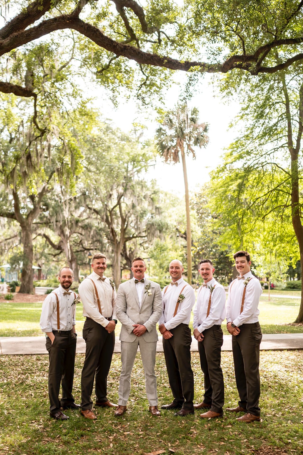 A wedding party stands in casual formal attire under Spanish moss-draped trees and palms in a sunlit Southern park setting.