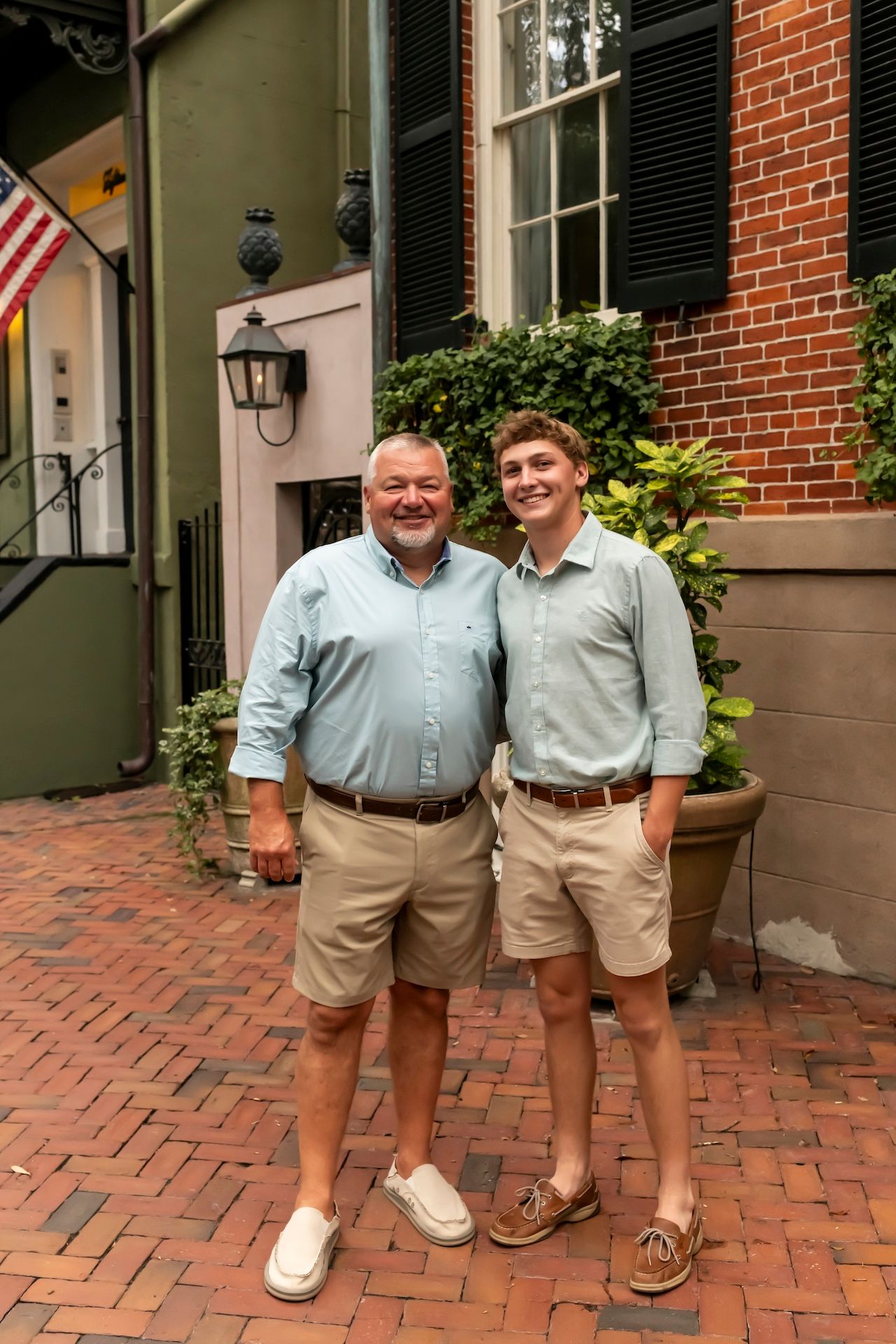 Two people in coordinating khaki shorts and collared shirts stand together on a brick sidewalk outside a historic building.