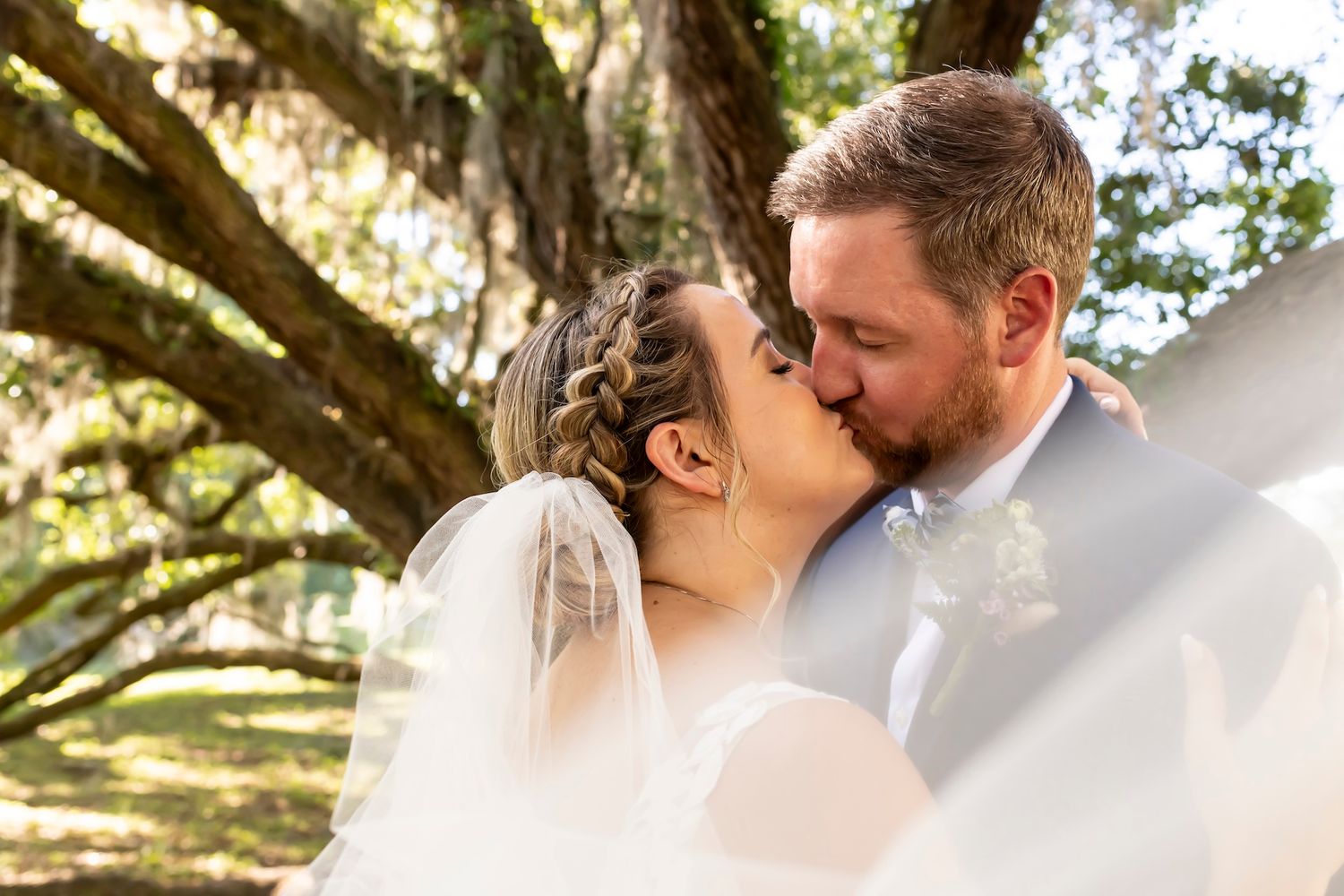 A romantic newlywed couple shares a tender kiss under sunlit oak branches during their outdoor wedding ceremony.
