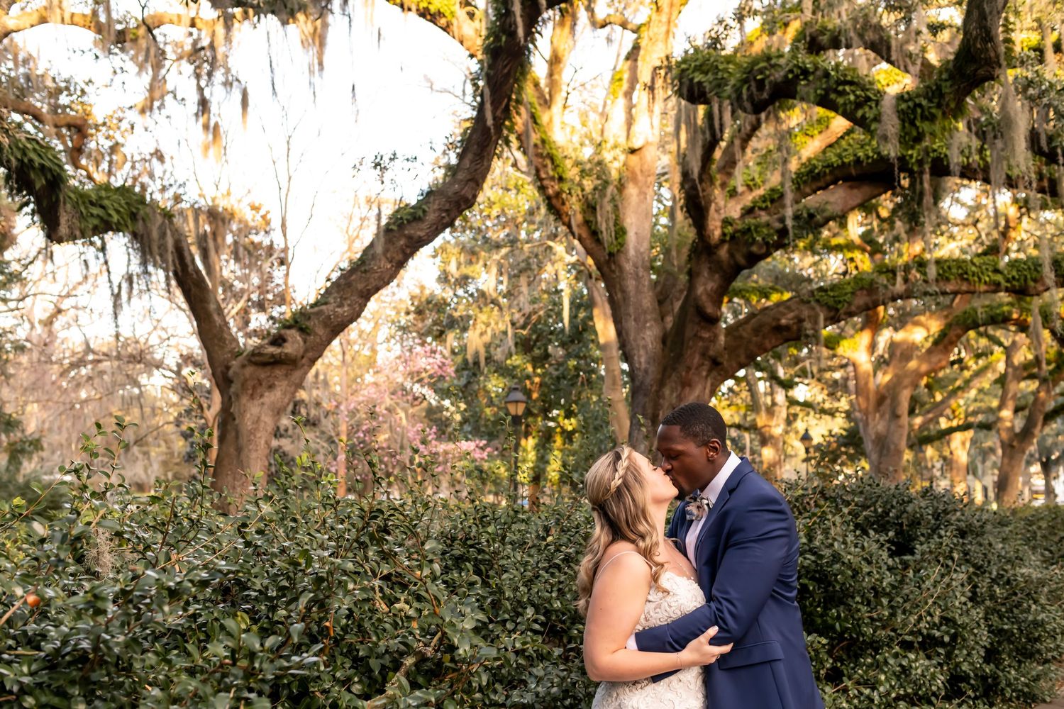 A romantic couple embraces under Spanish moss draped oak trees at sunset in a lush garden setting.